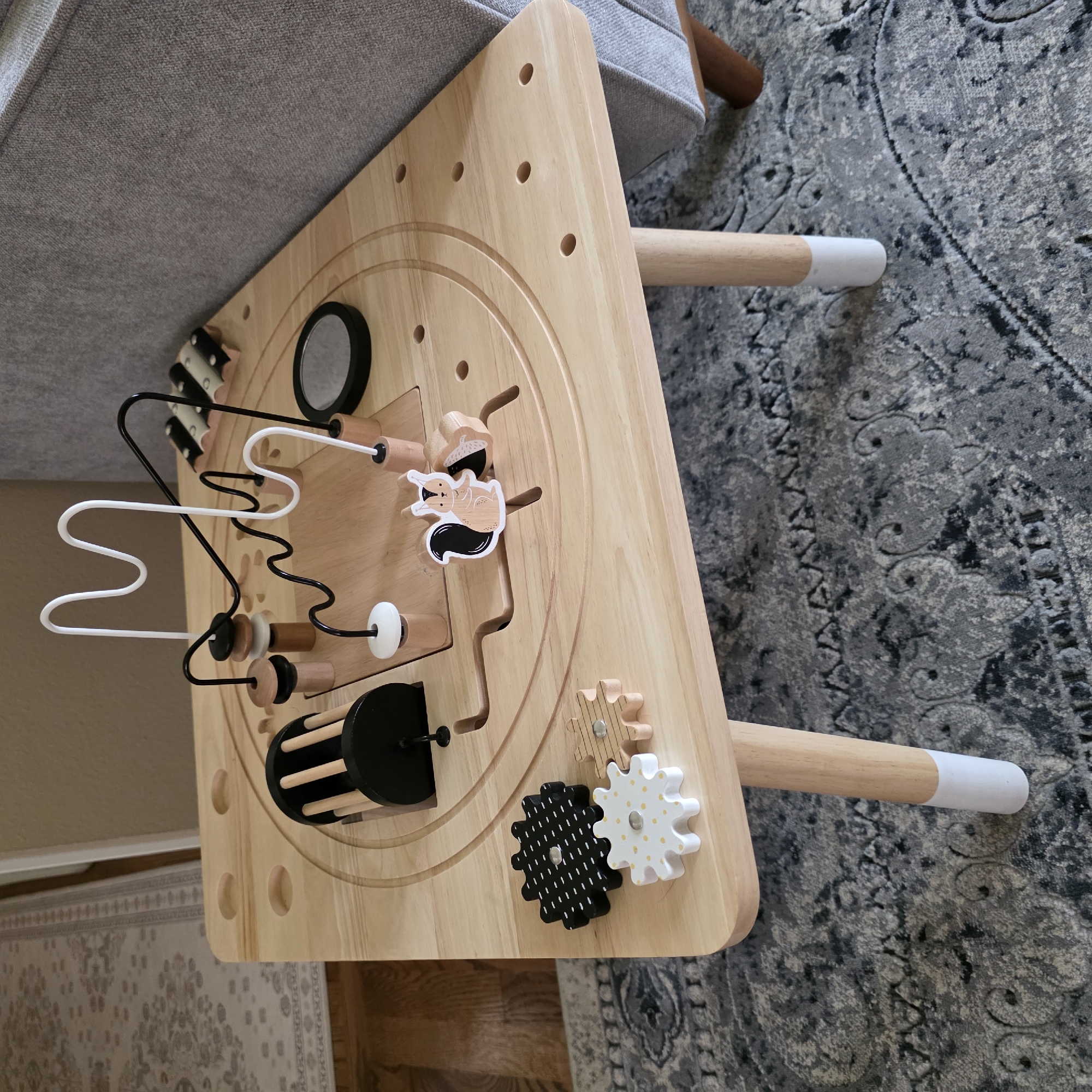 An indoor shot shows a wooden activity table with a bead maze, gears, and other interactive toys. The table is square with rounded corners and four wooden legs, two of which are visible and capped with white. The table's surface has various circular indentations and a central circular cutout containing the bead maze. A black and white squirrel sticker is placed on the table near the bead maze. To the left of the bead maze are three wooden gears, one black with white dots, one white with yellow dots, and one natural wood. The table sits on a gray and white patterned rug. In the background, a gray sofa is partially visible to the right, and a light-colored rug and wooden floor are visible to the left. The scene is well-lit with natural light. The location is not specified in the image.