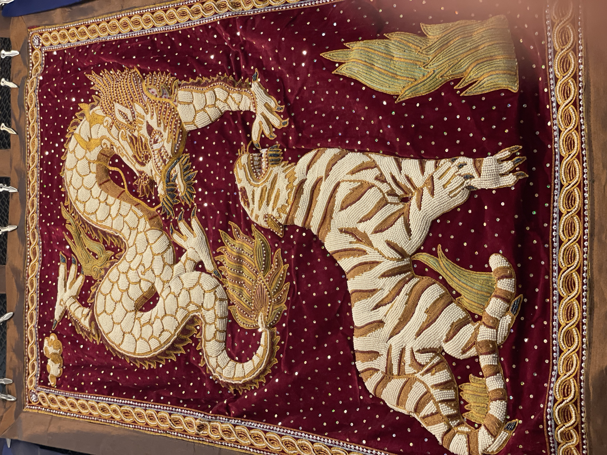 An ornate, beaded tapestry depicts a white and gold dragon coiled above a white and brown striped tiger on a deep red velvet background. The tapestry is adorned with small, shimmering sequins scattered across its surface. A decorative border frames the tapestry, featuring intricate gold and white beadwork. The tapestry is hung from metal clips against a dark background, with what appears to be part of a display stand visible at the top. The scene appears to be an indoor setting, possibly a museum or exhibition. The tapestry is likely a decorative art piece, with no people or specific actions depicted. The location is identified as Pattaya City, Thailand.