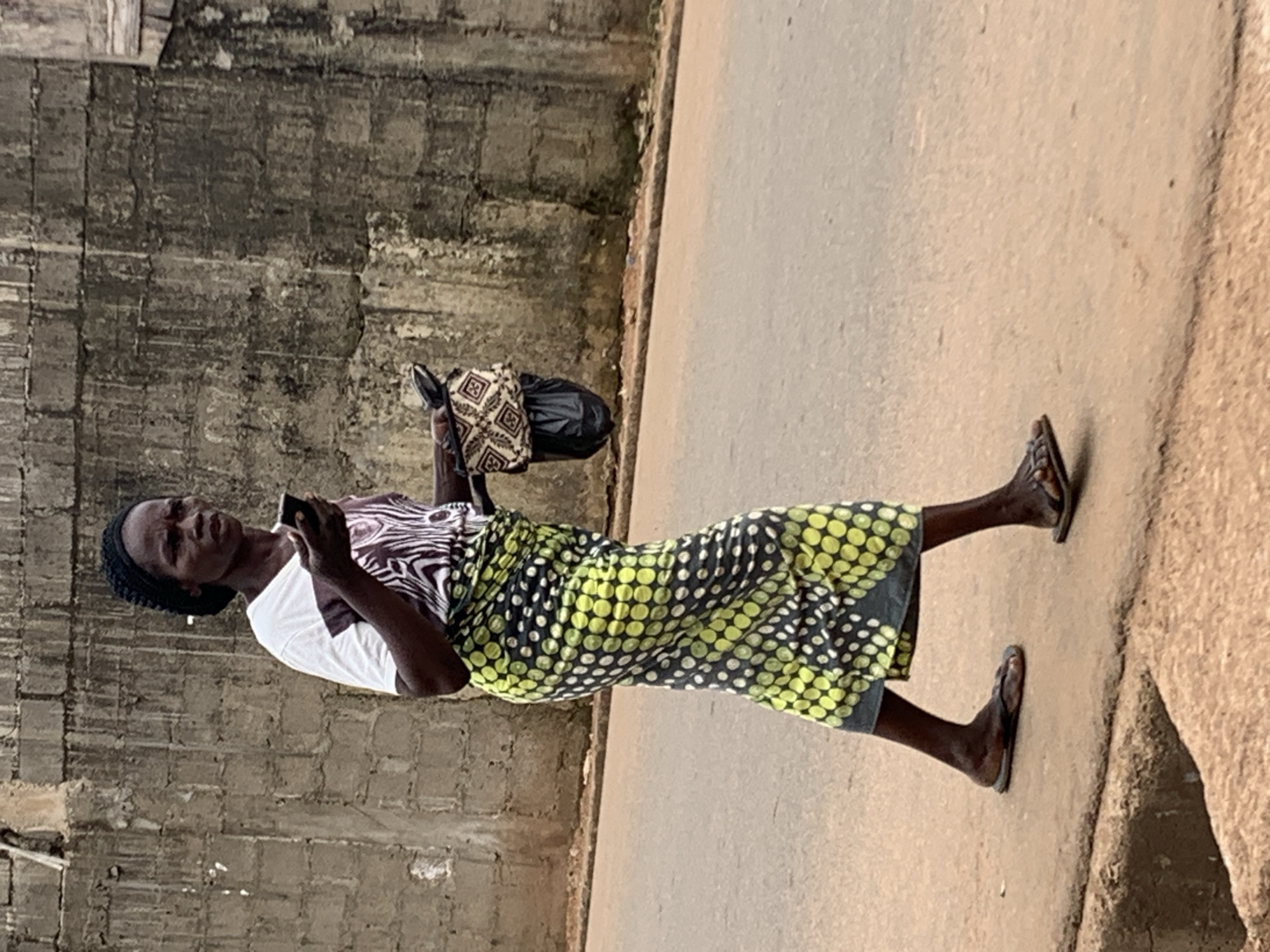 A woman is walking on a paved street in Akwanga, Nigeria. She is wearing a patterned skirt with green and black polka dots and a white t-shirt with a zebra-striped pattern on the front. She has a black head covering and is looking at a cell phone in her hands. She is also carrying a patterned bag in her other hand. She is wearing sandals. A textured, concrete wall runs along the left side of the frame. The street is on the right side of the frame.