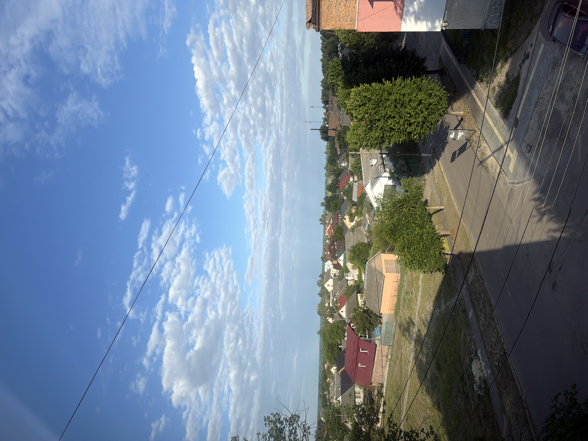 This is an eye-level, medium-distance, vertical shot of a residential area in Sosnivka, Ukraine, under a bright blue sky dotted with scattered white clouds. The scene depicts a mix of single-family homes with pitched roofs in various colors, including red, grey, and brown, interspersed with lush green trees. A paved road with visible tire marks runs through the middle ground, casting long shadows. Power lines crisscross the frame, originating from a multi-story brick building on the right side of the image, which features a pink and white facade. The foreground is dominated by the shadows of these power lines and the building. A dark-colored car is partially visible in the lower right corner. The overall environment suggests a tranquil, suburban setting during daylight hours.