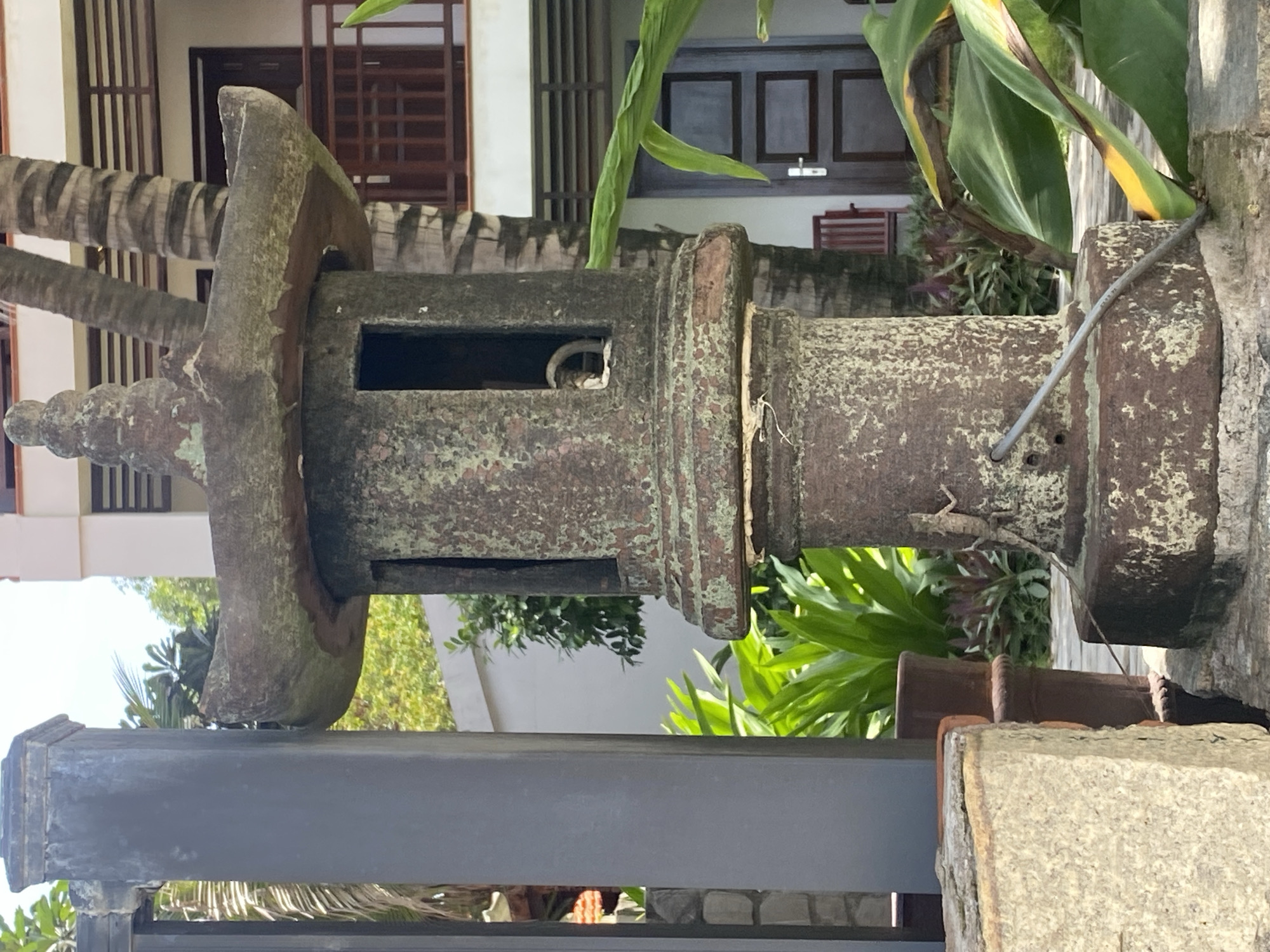 The image shows a weathered stone pagoda lantern in the foreground, with a light-colored stone pillar to its left and lush green foliage and a building in the background. A small lizard is visible on the base of the lantern. The lantern is cylindrical with decorative openings and a layered roof, covered in green moss and lichen, indicating age and exposure to the elements. Behind the lantern, a multi-story building with balconies and wooden doors is partially visible, surrounded by palm trees and tropical plants. The scene appears to be an outdoor setting, possibly a garden or courtyard of a resort or residential complex. The lighting suggests it is daytime, with bright sunlight casting shadows. The overall impression is one of tropical serenity and ancient charm.