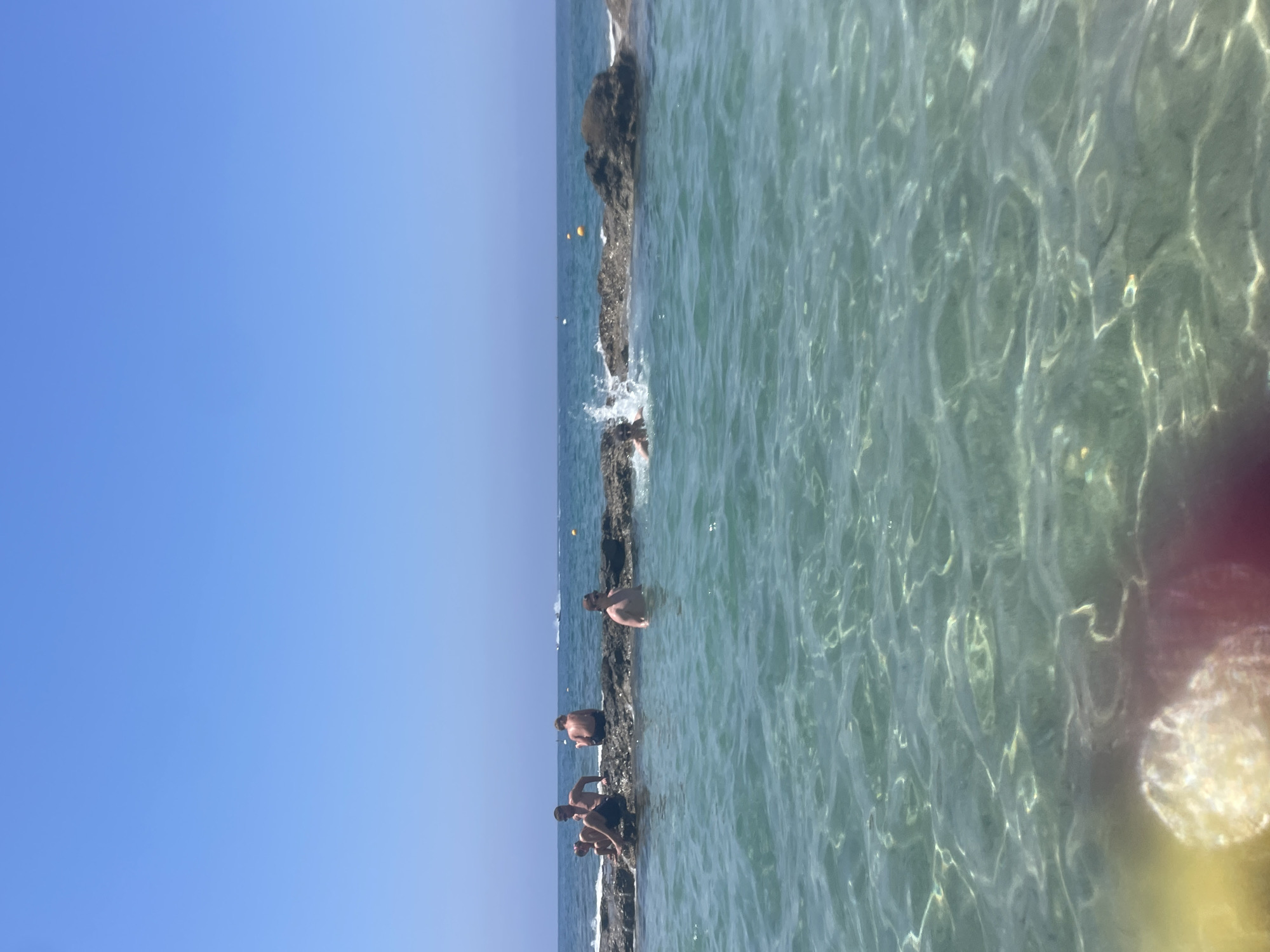 The image depicts a clear, sunny day at a coastal location, likely Paphos, Cyprus. In the foreground, the shallow, turquoise water is visible, with sunlight creating shimmering patterns on the sandy seabed. Several people are present, some seated on a rocky outcrop extending into the sea, while others are in the water. Two individuals are standing in the water near the rocks, and a third person is visible further out, splashing. In the background, the calm sea stretches to the horizon, meeting a clear blue sky. Buoys mark areas in the water, and a jet ski is visible in the distance. The overall scene conveys a relaxed atmosphere of a day spent at the beach or a natural rock pool.
