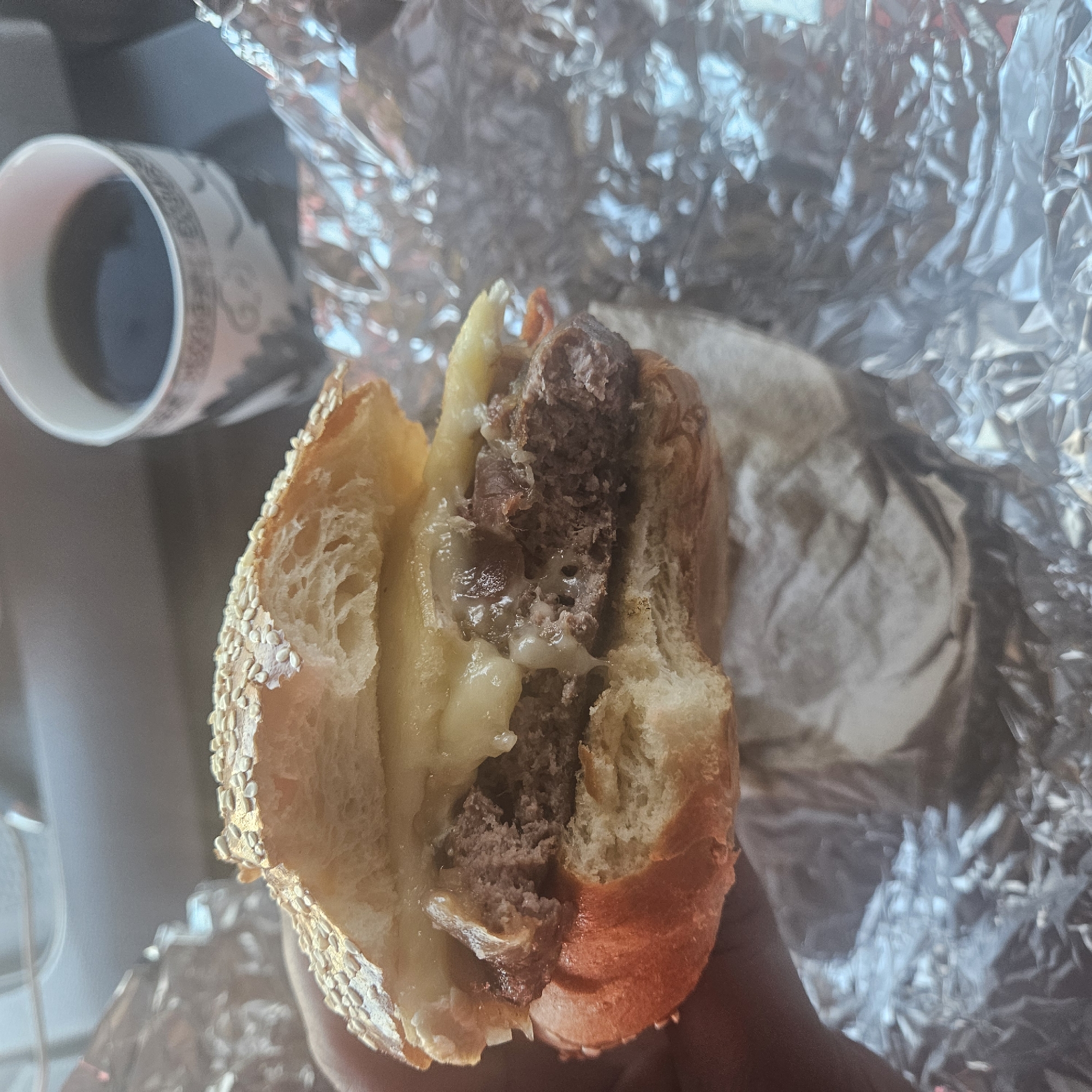 A partially eaten cheeseburger is held in someone's hand. The burger appears to be wrapped in foil. A coffee cup sits in the background. The scene is likely indoors. There is no specific information to confirm it is in Lleida, Spain.