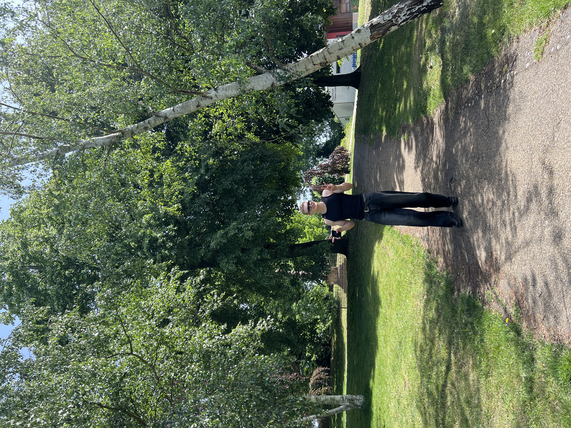 A figure stands on a gravel path in Christchurch Park, Ipswich, UK. The park is characterized by mature trees providing substantial shade. The figure, presumably male, is attired in dark trousers and a dark tank top. He wears sunglasses and a hat. The figure appears to be holding a phone. A birch tree stretches across the scene. The lighting indicates daylight. The scene includes a path and grassy areas. There is a fence on the park boundary.