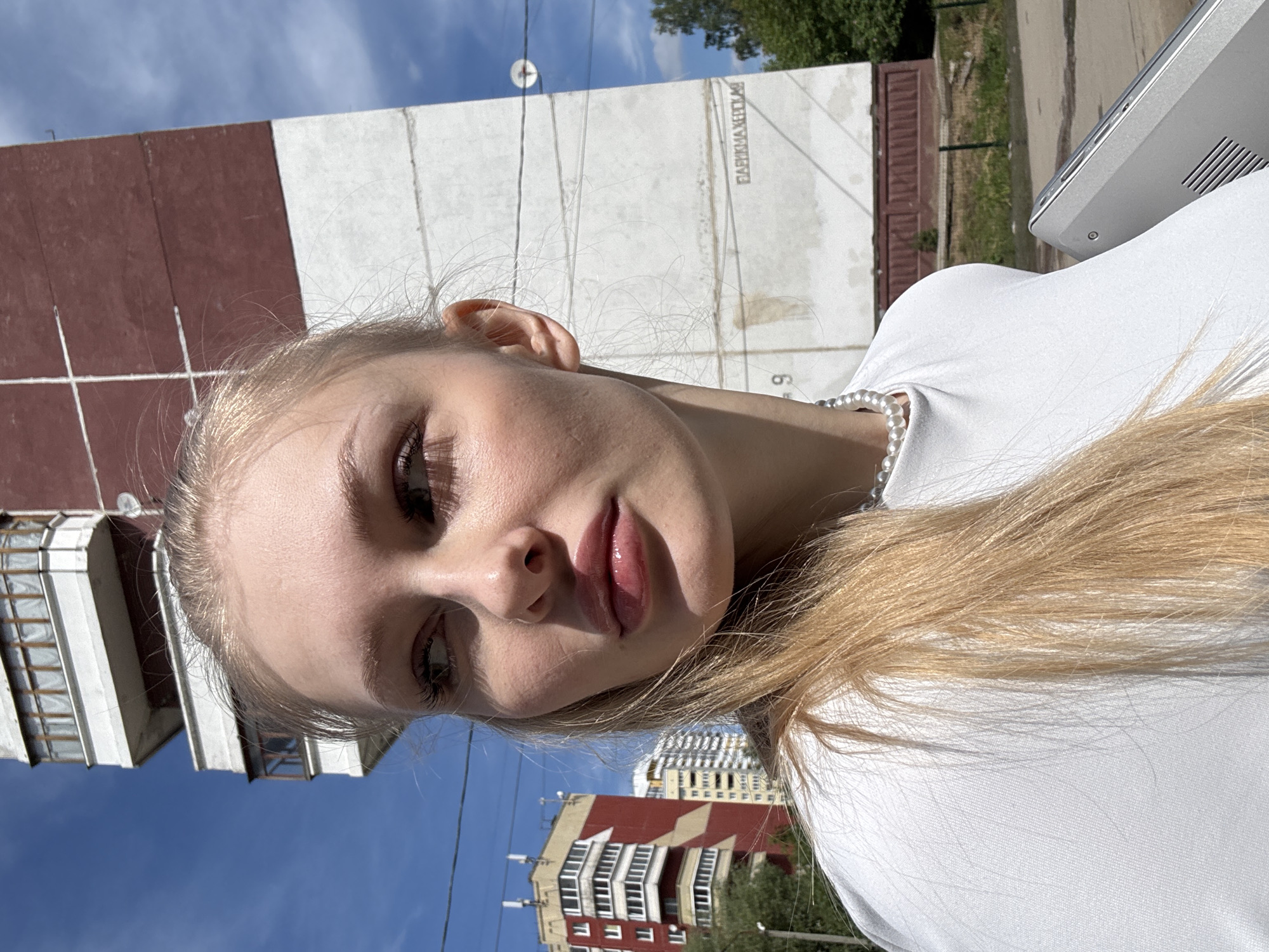 This close-up, angled outdoor portrait features a young blonde woman. She is wearing a white t-shirt and a pearl necklace, with a silver laptop or tablet partially visible on the right. Her gaze is directed slightly downwards and to the side, with a neutral expression and glossy lips.
The scene is set outdoors under a clear blue sky and bright sunlight, indicating daytime. In the background, urban buildings are visible. A prominent structure features two-tone red-brown and white sections. On a large white wall, likely part of a building, Cyrillic text clearly reads "ФАБРИКА ХЛЕБА" (FABRIKA KHLEBA), meaning "Bread Factory," with a faint number "9" visible below it. These details, combined with the provided location context, suggest the image was taken in Perm, Russia, on a sunny day.