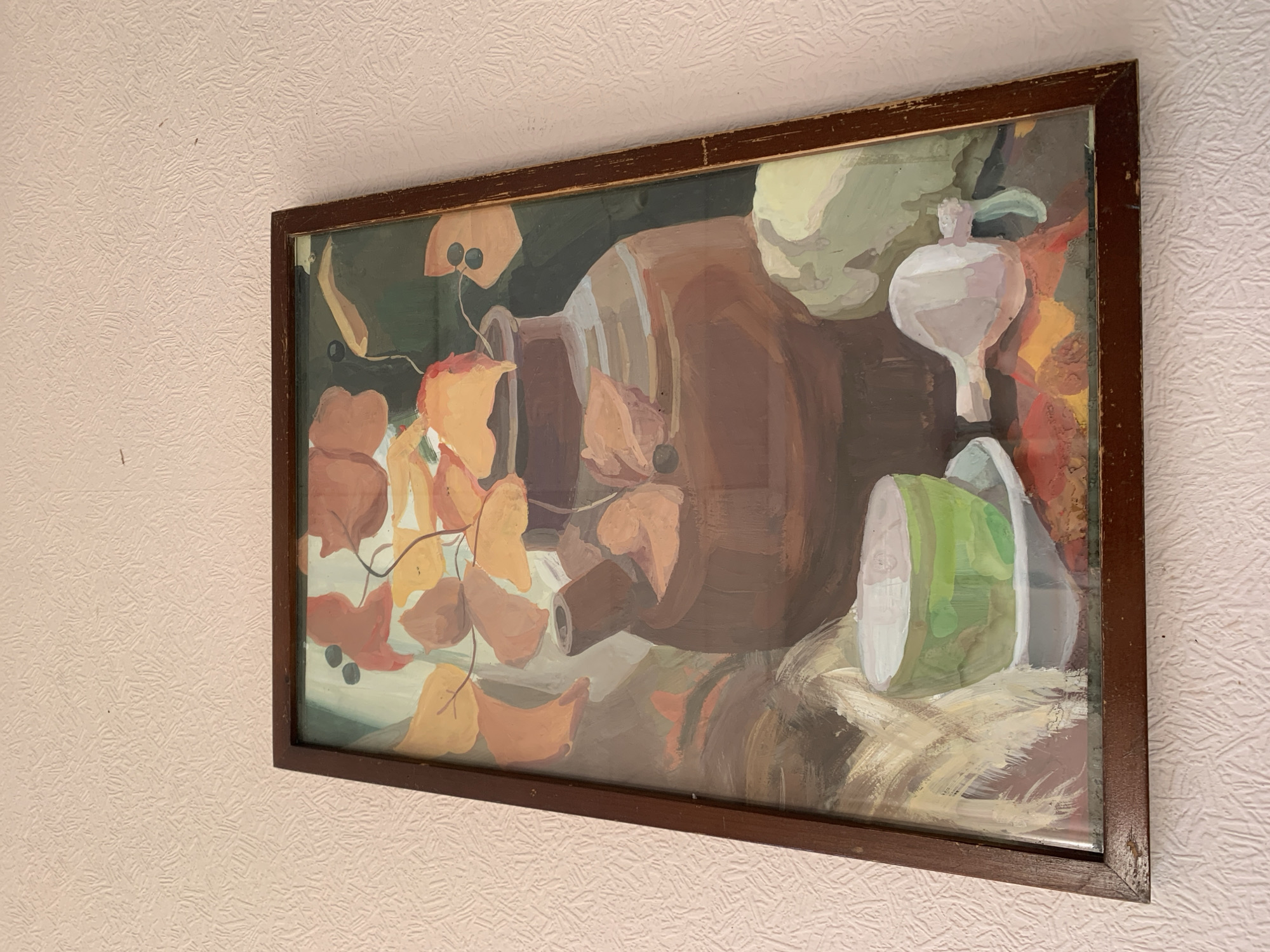 The image shows a framed painting hanging on a light pink textured wall. The painting depicts a still life with a brown ceramic pitcher, autumn leaves, a white onion or garlic, and a green and white container. The painting is done in a somewhat impressionistic style with broad brushstrokes. The frame is dark brown and appears to be aged. The location is not evident from the provided image. No people, actions, or interactions are depicted.