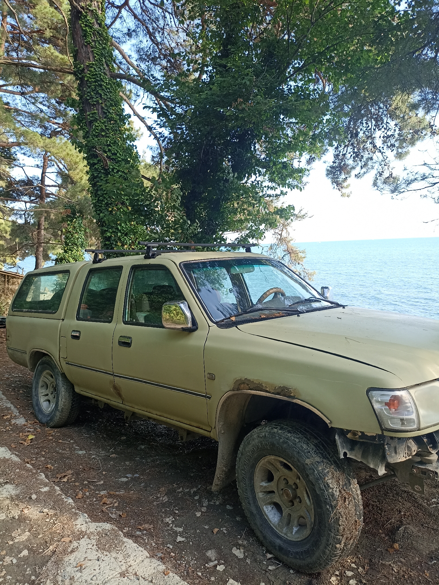 This media file shows a pale green, four-door pickup truck with a camper shell, parked outdoors on a dirt and gravel path next to a large body of water, likely the sea. The truck appears to be older and somewhat weathered, with visible rust on the wheel arches and some damage to the front bumper. It has a roof rack installed. The scene is set amidst trees, with a tall tree heavily covered in ivy prominently featured on the left side of the image. The sky is blue with some clouds, and the water is a calm blue. The lighting suggests it is daytime, possibly late afternoon, given the quality of the light and the shadows. The overall impression is one of a remote or somewhat secluded location, possibly a campsite or a natural area. There is no visible text in the image. The setting, with the combination of trees, dirt path, and the sea, along with the mention of Sochi, Russia, suggests a coastal, natural environment. The truck's condition might imply it's being used for outdoor activities or is perhaps abandoned or in a state of disrepair in a scenic location. No people are visible in the image.