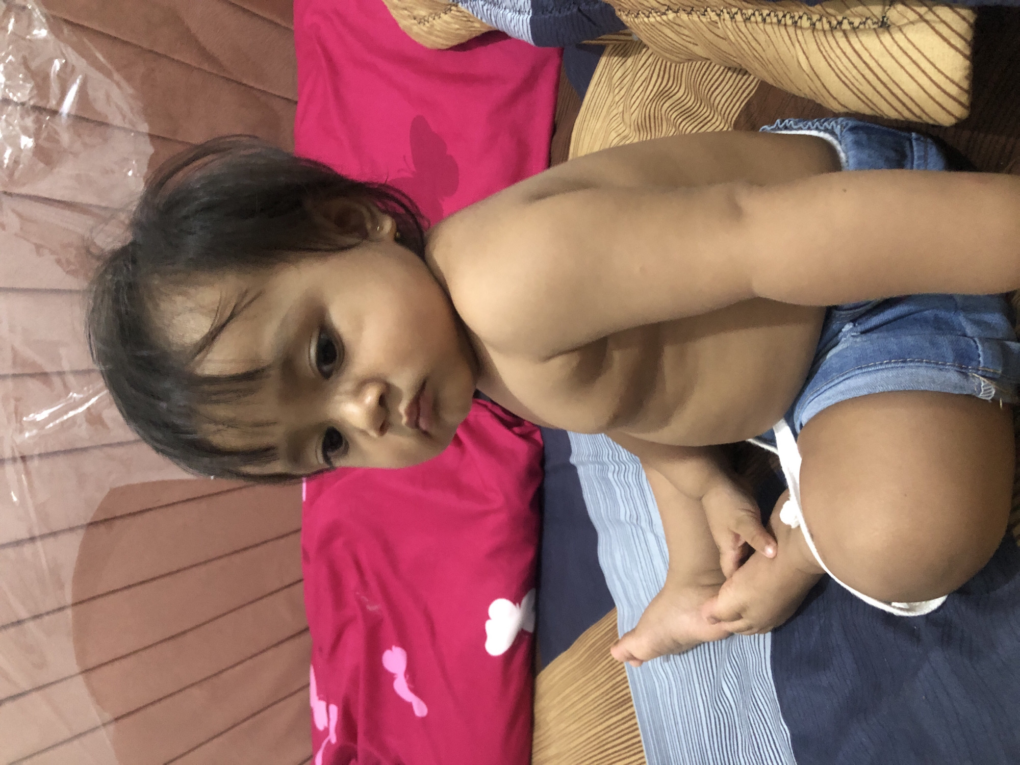 A young child, likely a toddler, is depicted in a close-up, top-down shot. The child has dark hair and is looking directly towards the camera with a neutral, slightly pensive expression. They are wearing only denim shorts, leaving their torso bare, and are positioned in a relaxed manner on what appears to be a bed or soft surface.
The setting consists of various soft furnishings. To the child's left is a vibrant pink pillow or blanket adorned with a white butterfly design. Below the child's body, and extending to the right, are striped blankets or sheets in shades of blue, light brown, and dark brown. A clear, crinkled plastic sheet or cover is visible on the surface to the upper left of the child, creating reflections. The lighting suggests an indoor setting, likely during the daytime. There is no visible text in the image. The scene takes place in Lhokseumawe, Indonesia.