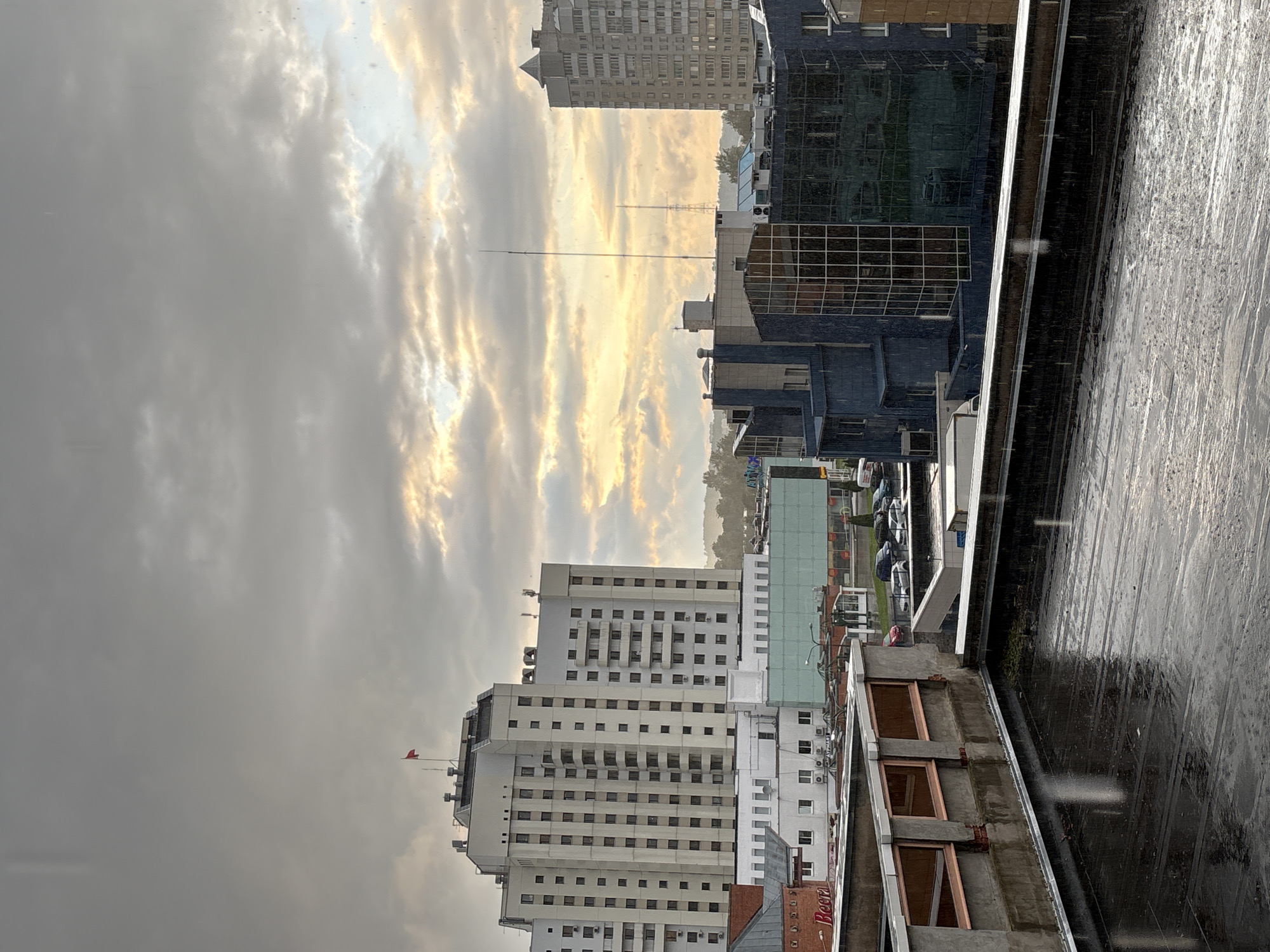 This is a high-angle, outdoor, eye-level shot of a city skyline on a cloudy, rainy day. In the foreground, a wet, dark-colored flat rooftop is visible, with reflections of the sky on its surface. A low, concrete wall with rectangular, orange-tinted windows runs along the edge of the rooftop. Beyond the rooftop, several multi-story buildings are visible. The largest building, a pale beige high-rise with numerous windows, is centrally located and has a red flag on its rooftop. To the right of this building are a series of dark blue, modern-looking buildings with glass facades. Further to the right, a tall, grey residential building stands. In the mid-ground, streets with parked cars are visible, and some of the buildings have signage. The sky is overcast with pockets of bright, yellowish light peeking through the clouds, suggesting the sun is low in the sky. The overall impression is of a city during a rain shower with diffused daylight. The specific location is Vitsebsk, Belarus, as indicated by the presence of the "Beera" store signage, a known establishment in Vitsebsk.