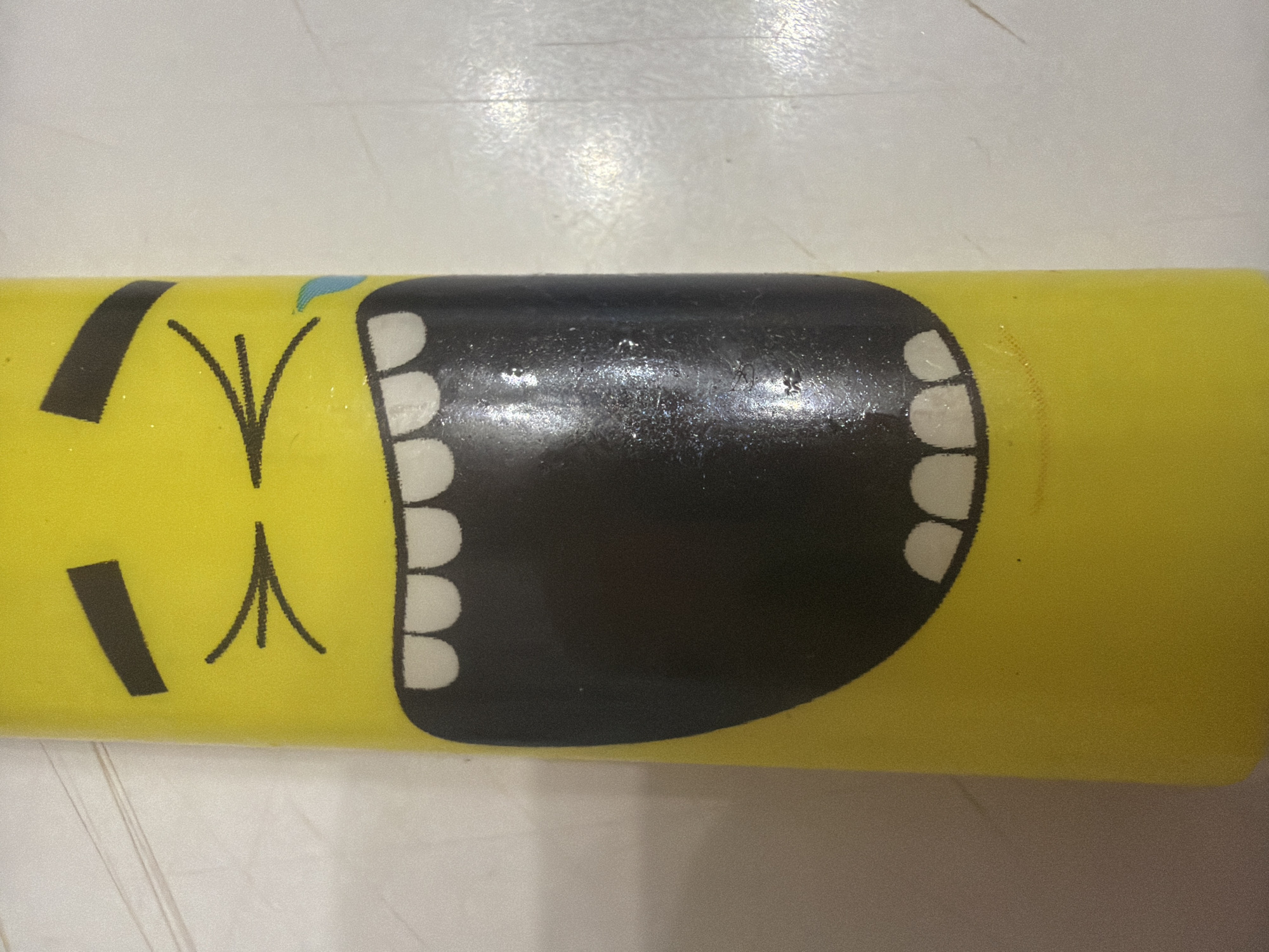 The image shows a close-up of a yellow cylindrical object with a cartoonish face printed on it. The face has black eyebrows, closed eyes that appear to be squinting, and a large open mouth with white teeth. A small blue teardrop is depicted near the right eye. The object is positioned on a light-colored, textured surface, possibly a tabletop, with some faint lines visible. The lighting appears to be overhead, creating some reflections on the surface of the object. There is no indication of the city of Sochi, Russia, in the image or its context.