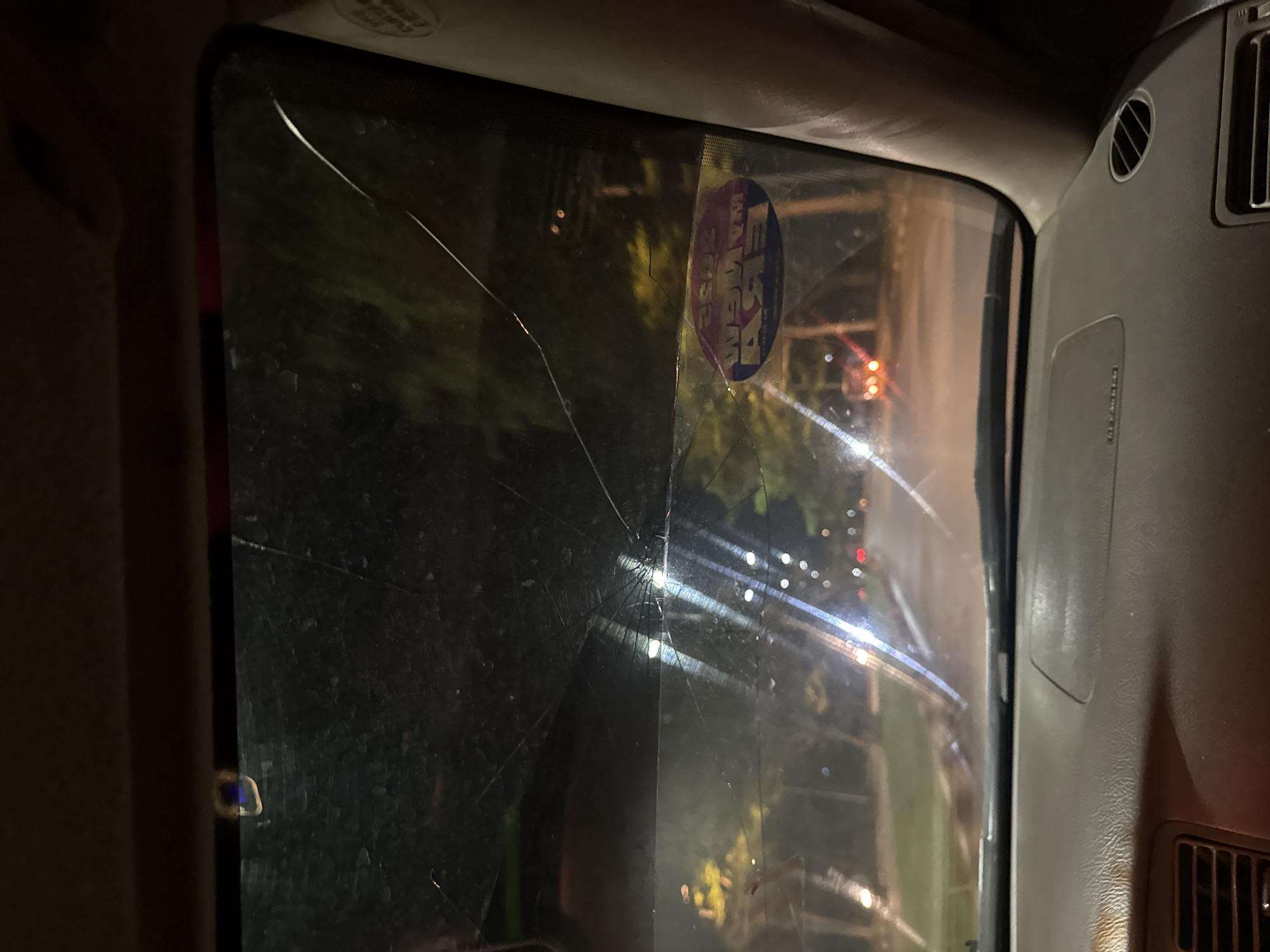 The image shows a cracked car window, possibly a windshield, viewed from the inside of the vehicle. The cracks originate from a central point, radiating outward. A round sticker is affixed to the window, obscuring the view. Outside the window, blurred lights and vague outlines of trees suggest a nighttime setting. The interior of the car is visible, with a dashboard and air vents. There is no evidence to confirm the location is Abuja, Nigeria.