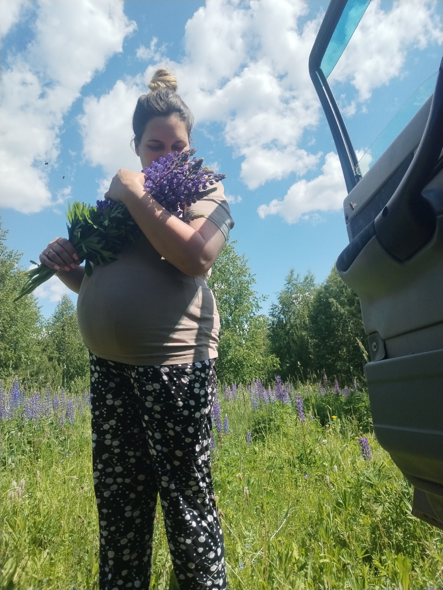 A pregnant woman with her hair in a bun is standing in a field of purple lupine flowers under a blue sky with white clouds. She is wearing a light brown t-shirt and black pants with a white floral pattern. She is holding a bouquet of purple lupine flowers and green leaves. To the right of the image, a portion of a vehicle's door and interior is visible. The scene appears to be a rural outdoor setting, likely in or around Zipunovo, Russia.