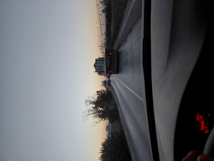 A view from inside a vehicle shows a highway at dusk. A multi-level trailer is driving in front. The sky is a gradient of pale blue to orange at the horizon. Trees and vegetation line the side of the road. A building is visible in the distance. Part of the car's interior is in the foreground, including the dashboard and steering wheel. The speedometer displays red numbers.