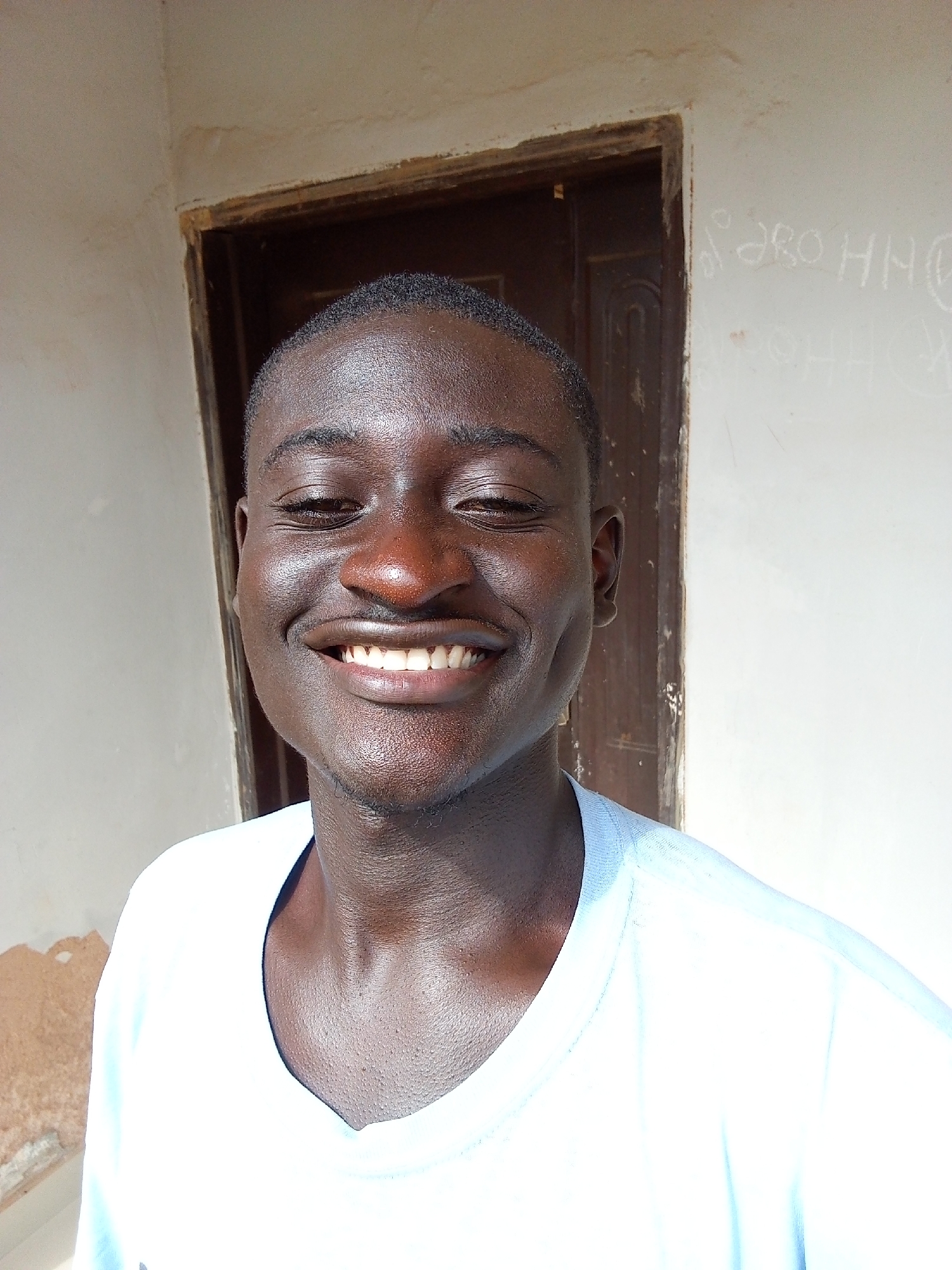 A young Black man is smiling directly at the camera. He has short black hair, dark brown skin, and white teeth. He is wearing a white t-shirt. Behind him is a brown wooden door and a white wall. The wall has faded writing on it. The picture appears to be taken outdoors in daylight. While the exact location within Bukuru cannot be determined from the image, the image suggests a residential area due to the presence of a door and wall.