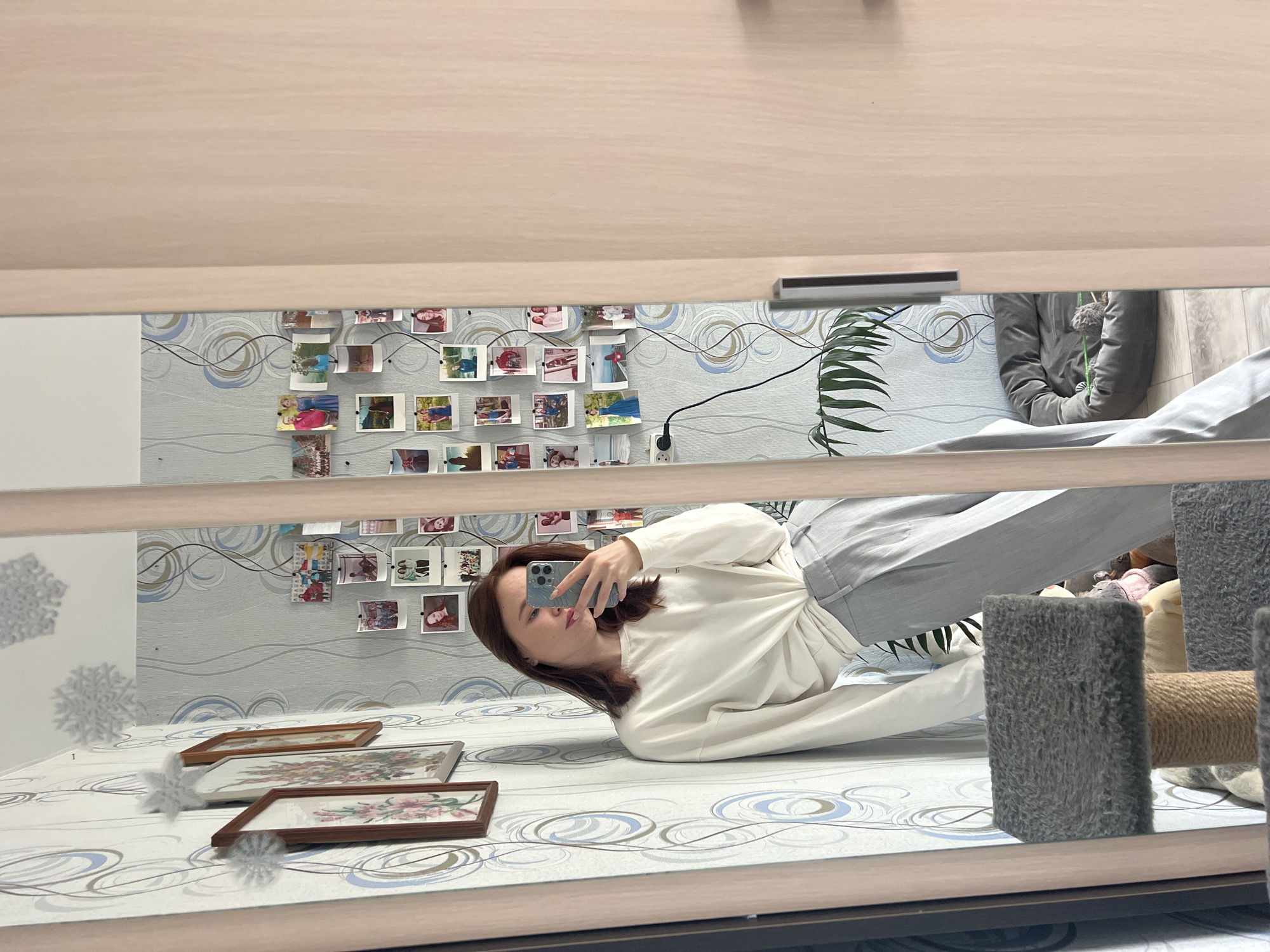 The image captures a selfie reflection of a young woman in a large, vertically oriented mirror, which appears to be part of a light-colored wooden wardrobe or closet door. The entire image is rotated 90 degrees clockwise.
**Subjects:** The primary subject is a young woman, likely in her 20s or early 30s, with medium-length brown hair. She is seen in a full-body reflection. A second person is partially visible in the background reflection, wearing a grey padded jacket.
**Objects:** The woman is holding a light blue smartphone (resembling an iPhone 13 or 14 Pro model) to take the selfie. She is dressed in a cream-colored long-sleeved top and light grey trousers. The mirror reflects a room decorated with light blue wallpaper featuring subtle grey and blue swirling patterns. Adorning the wall are numerous small, square-shaped photographs strung together with a dark wire. On the patterned surface where the woman is lying (likely a bed or sofa), three framed pictures, possibly cross-stitch art depicting floral arrangements, are displayed. Further into the reflected room, a cat scratching post with grey carpeted and natural fiber sections is visible. Other objects include a potted green plant with long leaves, an electrical outlet with a black cable plugged in, and decorative white snowflake cutouts adhered to the lower-left corner of the mirror surface.
**Activity or event taking place:** The woman is actively taking a selfie of herself reflected in the mirror. She is lying or reclining on a surface covered with a white sheet or blanket featuring blue and grey circular patterns, and she holds her phone up to capture the image.
**Notable details:** The woman has a neutral to slightly contemplative expression. The lighting in the room appears to be soft and ambient, suggesting daytime indoors. The style of the wallpaper and furniture indicates a residential interior. The decorative snowflakes on the mirror could hint at a winter season or a permanent festive decoration. There is no visible text in the image.
**Location:** Chaikovsky, Russia.
