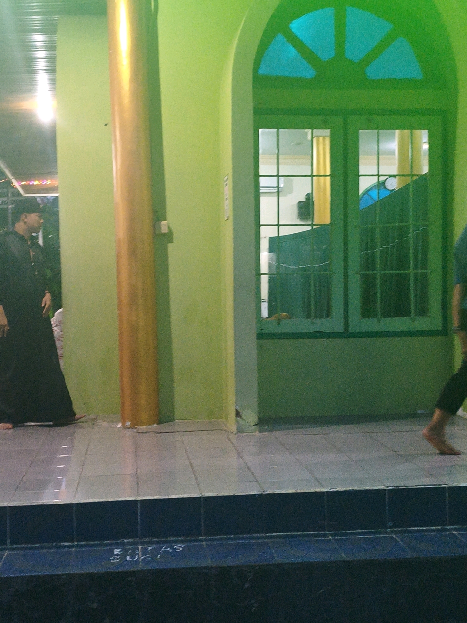 The image depicts an outdoor scene, likely a walkway or entrance to a building, illuminated by a greenish light. In the foreground, a man dressed in dark, traditional attire, possibly a thobe and kufi, is seen walking away from the viewer towards the left side of the frame. His right foot is visible, and he appears to be barefoot.
To the right of the man, a thick, golden-colored cylindrical pillar stands vertically. Behind the pillar, a large, arched window with green frames and divided panes is visible. The window appears to be covered with a dark material, and through it, faint reflections and objects within the building can be discerned, including hints of yellow pillars and blue structures.
The ground is tiled with light gray square tiles, leading up to a dark blue tiled step or ledge in the foreground. White markings, possibly painted lines, are visible on the gray tiles. The overall impression is of an evening or dimly lit environment. The specific location in Samarinda, Indonesia, is not explicitly discernible from the visual elements alone, but the architectural style and the man's attire might suggest a cultural context relevant to the region.
