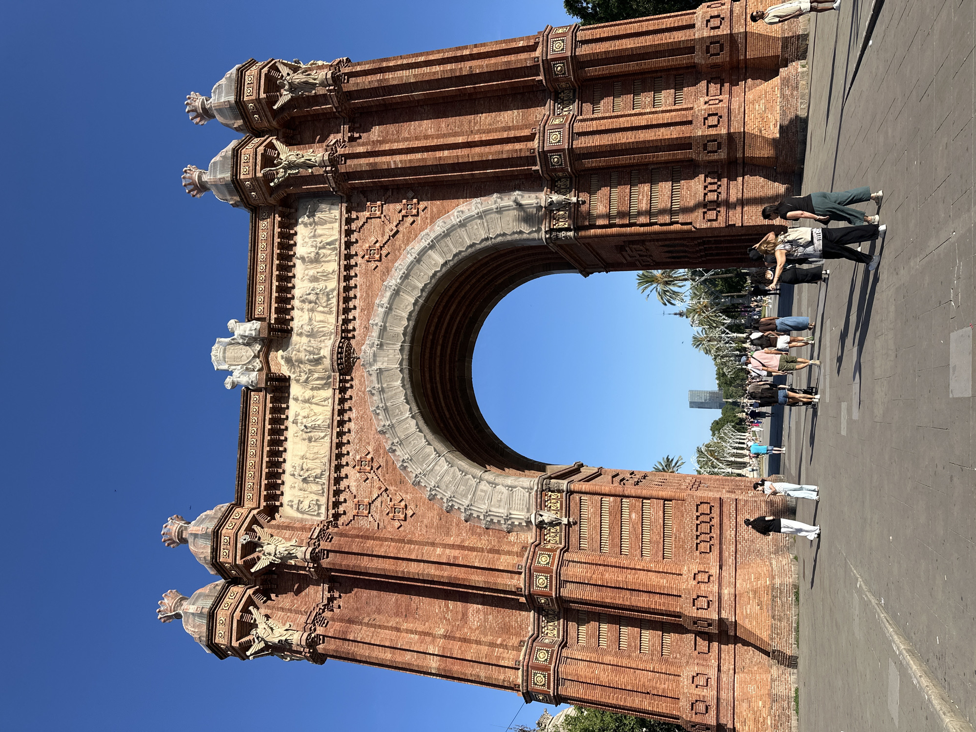 The image depicts the Arc de Triomf in Barcelona, Spain, viewed head-on from a slight low-angle perspective on a clear day. The monumental archway, constructed predominantly of reddish-brown brick, features intricate architectural details including sculpted figures, decorative friezes, and four prominent cupola-like structures on its upper corners. The central arch is adorned with lighter-colored stonework. The sky above is a uniform, vivid blue with no clouds, and small dark specks, likely birds, are visible in the upper right quadrant.
A wide, paved promenade extends in front of the arch, made of light-colored flagstones. Numerous individuals are present on the promenade, engaging in various activities such as walking, standing, and conversing. Most are dressed in casual, warm-weather attire, including t-shirts, shorts, and light trousers. On the right side of the image, a person in dark pants and a white top is seen with an arm extended, possibly taking a photograph or gesturing. Near them, a stroller is visible. On the left side, two individuals are standing and interacting. Strong sunlight illuminates the scene, casting distinct shadows of the people and the archway onto the pavement. Through the archway, a wide avenue lined with palm trees is visible, leading to a distant modern skyscraper in the center of the frame.