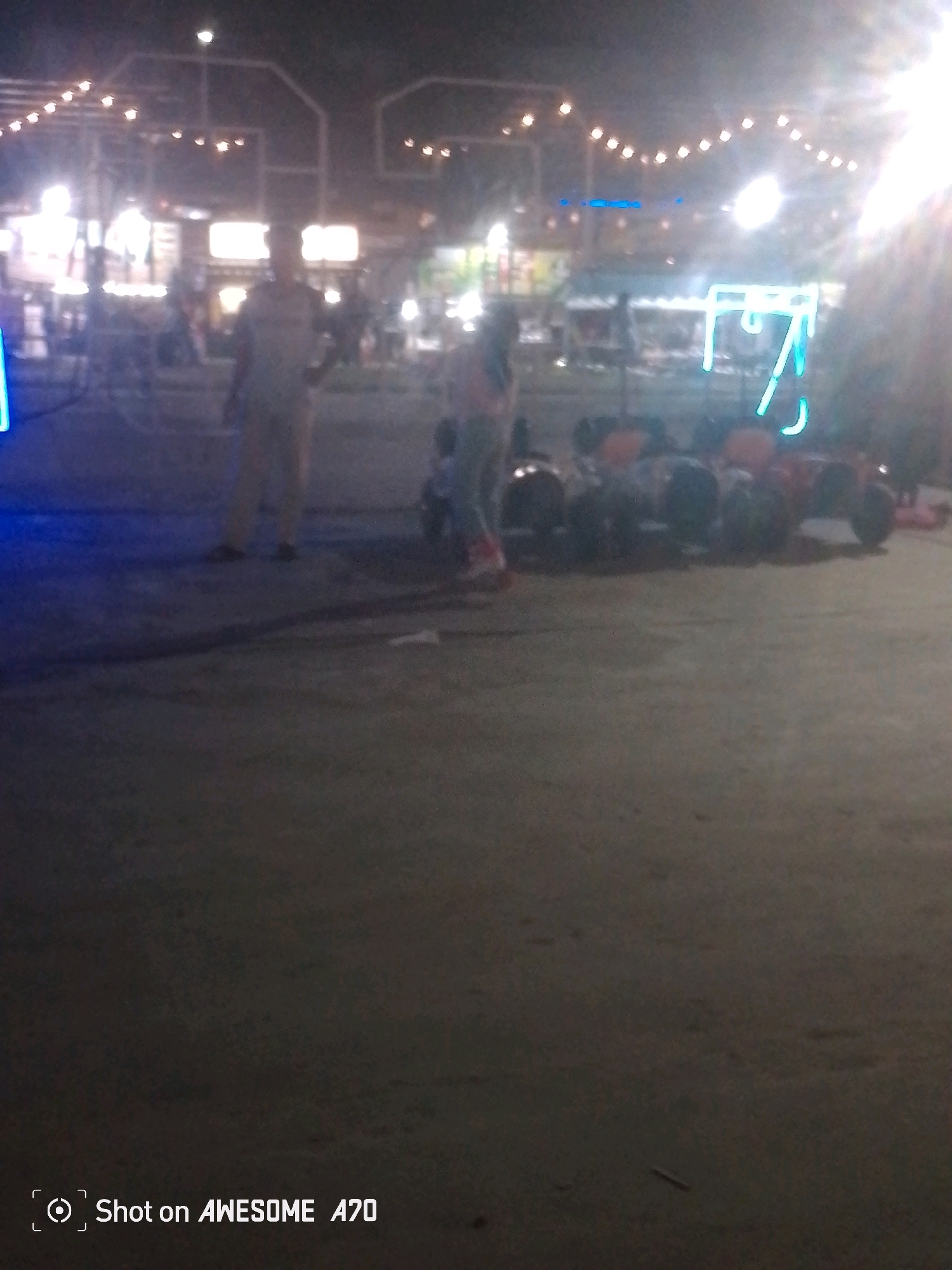 In the city of Batam, Indonesia, at an outdoor establishment illuminated by string lights and neon signage, two individuals are present. One person, a male, is standing in the foreground. Behind him, a second individual, a female, is seen near a row of parked vehicles, possibly small go-karts or ATVs. The vehicles appear to be for recreational use, with some showing orange accents. The ground is a paved surface. The overall ambiance suggests a nighttime entertainment or recreational area.