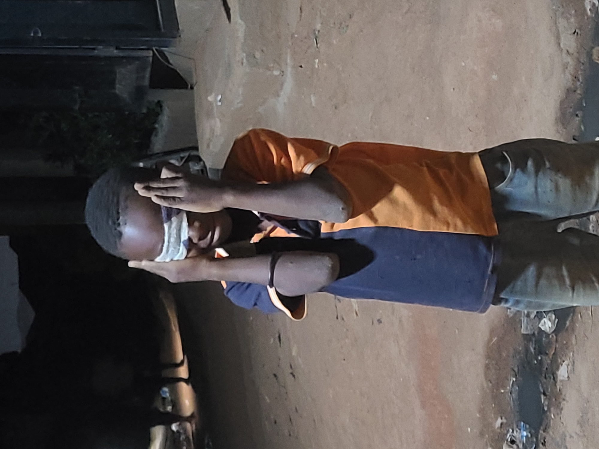 An individual is outdoors in an urban location. The individual is wearing light-colored trousers, a two-tone shirt of yellow and dark blue, and has a white bandage covering their eyes, held in place by their hands. The setting appears to be a street with an uneven surface, possibly in Jos, Nigeria. Shadows are present, suggesting a low light or nighttime setting. There are some objects or trash on the ground near a dark or dirty area. Part of a building is visible in the background.