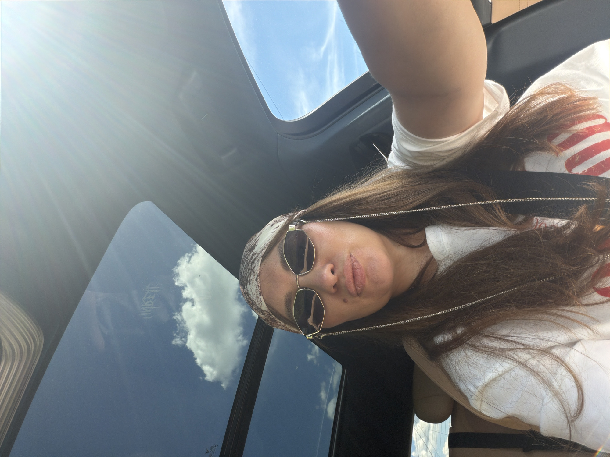 A woman is taking a selfie while in a car with a sunroof open. The sky is visible through the sunroof, with some clouds and sunrays. The woman is wearing sunglasses, a bandana on her head, and a white t-shirt with red stripes. She is looking at the camera and pouting her lips. The interior of the car is visible, including a leather seat and part of the dashboard. The location is not identifiable as Malinovka, Russia, from the provided image.