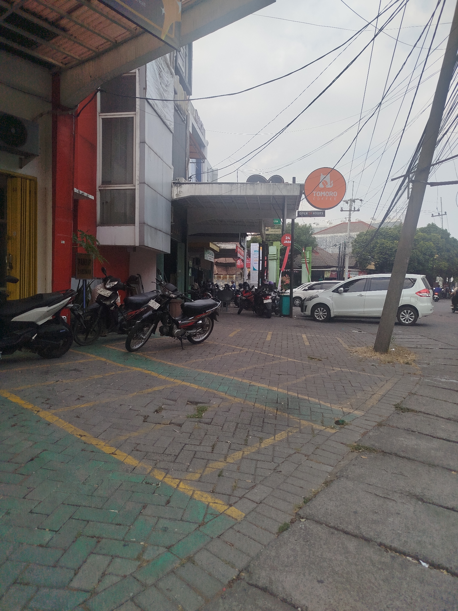 The image captures a street scene in Yosowilangun, Indonesia, on an overcast day. In the foreground, a paved area is marked with yellow and green parking lines, indicating a parking lot. Several motorcycles are parked in this area, mostly to the left and center of the frame. A white minivan is parked on the right side of the street, facing away from the camera. Behind the minivan, a utility pole with numerous electrical wires stretches across the frame.
A building with a red facade and a modern gray extension occupies the left and center background. A circular orange sign with a white logo and the text "TOMORO COFFEE" is visible above a covered walkway. Further back, more buildings and trees can be seen, suggesting a commercial or mixed-use area within the town. The overall environment appears to be a typical urban setting with a mix of commercial establishments and street infrastructure. There are no people clearly visible or actively engaged in any significant action within the frame. The scene is static, focusing on the arrangement of vehicles, buildings, and overhead wires.