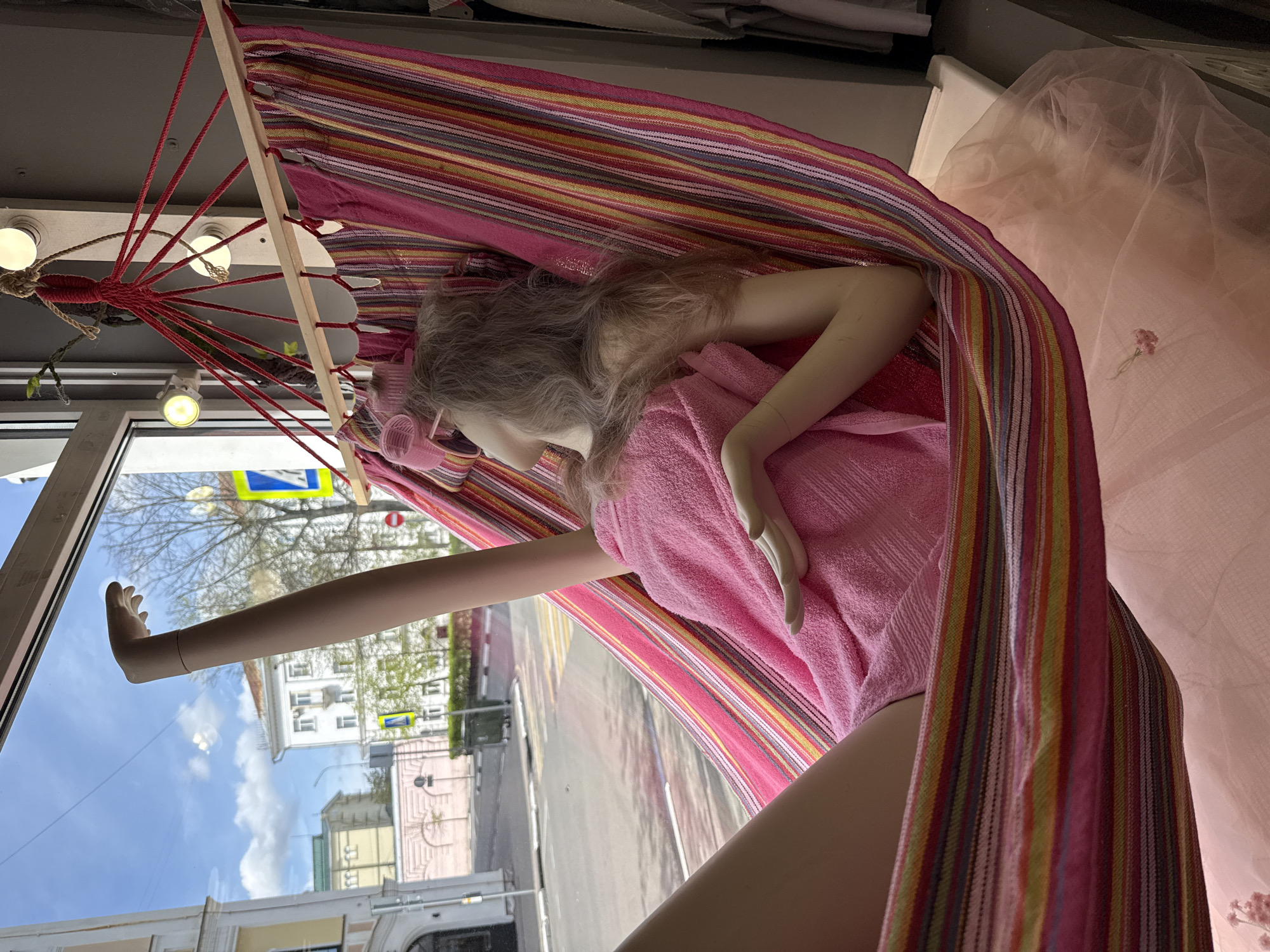 A window display in Заволжье, Russia, features a mannequin reclining in a striped hammock. The mannequin wears a pink robe and has hair rollers in its hair. The hammock is suspended by a wooden frame and red ropes. Outside the window, buildings and a street scene are visible, reflecting the sky.