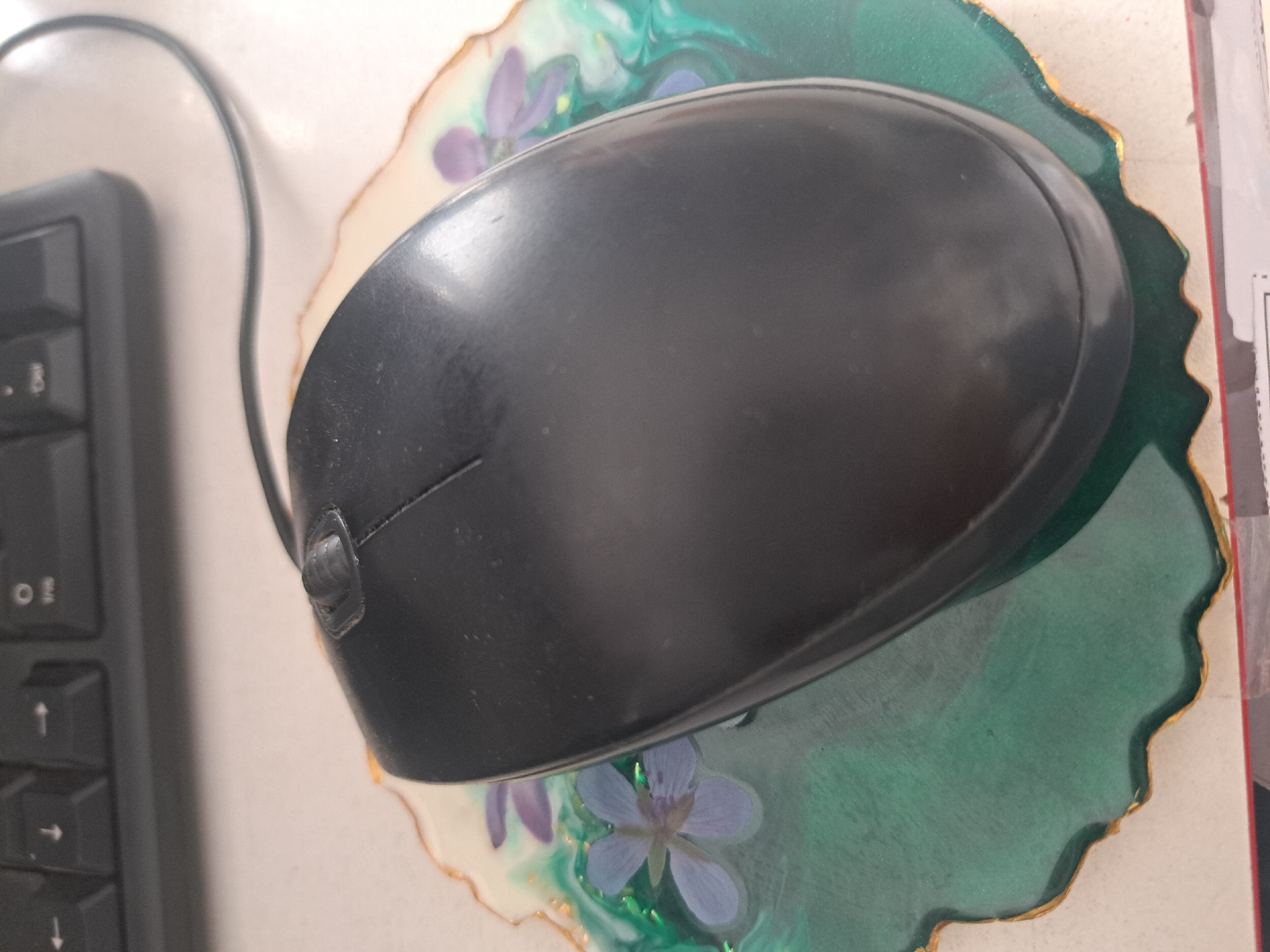 A black computer mouse sits on a green and white floral-themed mousepad, placed next to a black keyboard on a white surface. There is nothing in the image to indicate that this image was taken in the city of Leningradskaya, Russia.