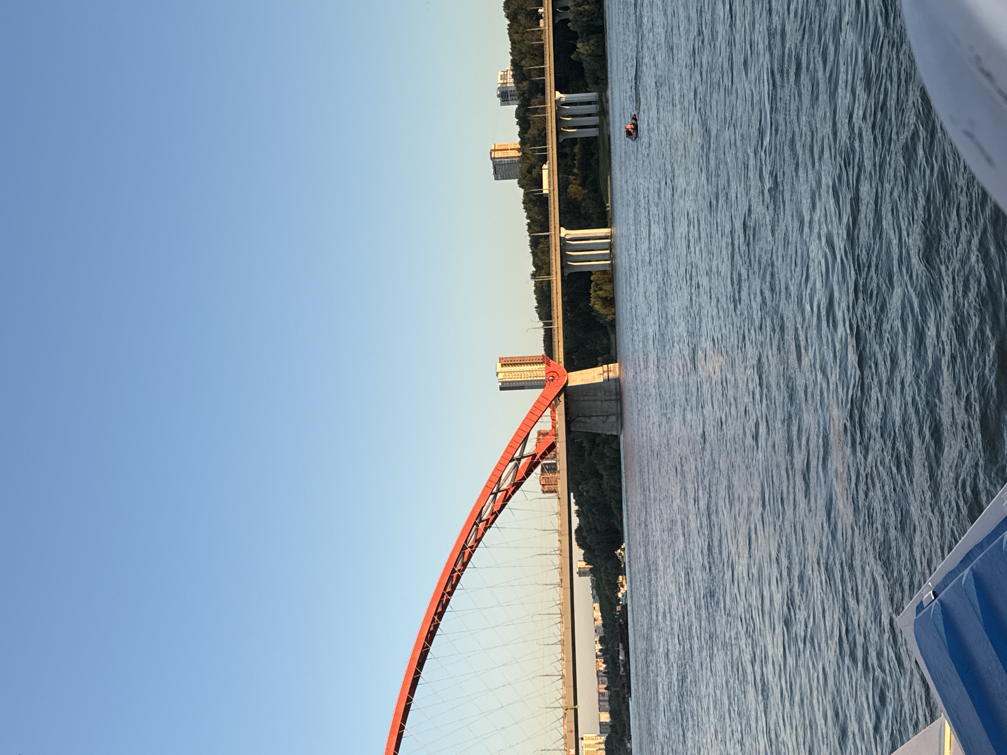 This is a photograph taken from a body of water, likely a river, showing a large red suspension bridge in the foreground, spanning across the water. The bridge has a distinct arch and is supported by piers. In the background, another, straighter bridge with multiple concrete piers is visible, carrying vehicular traffic. Beyond the bridges, a treeline with scattered buildings, including tall apartment complexes, can be seen along the riverbanks. The sky is clear and blue, indicating fair weather, possibly during sunset due to the warm lighting on the bridge. There is a small boat with two people in it on the water in the middle ground. The foreground also shows a portion of another boat or watercraft. The overall scene depicts a cityscape with significant infrastructure and natural elements. The bridge in the foreground is the Bugrinsky Bridge, a prominent landmark in Novosibirsk, Russia.