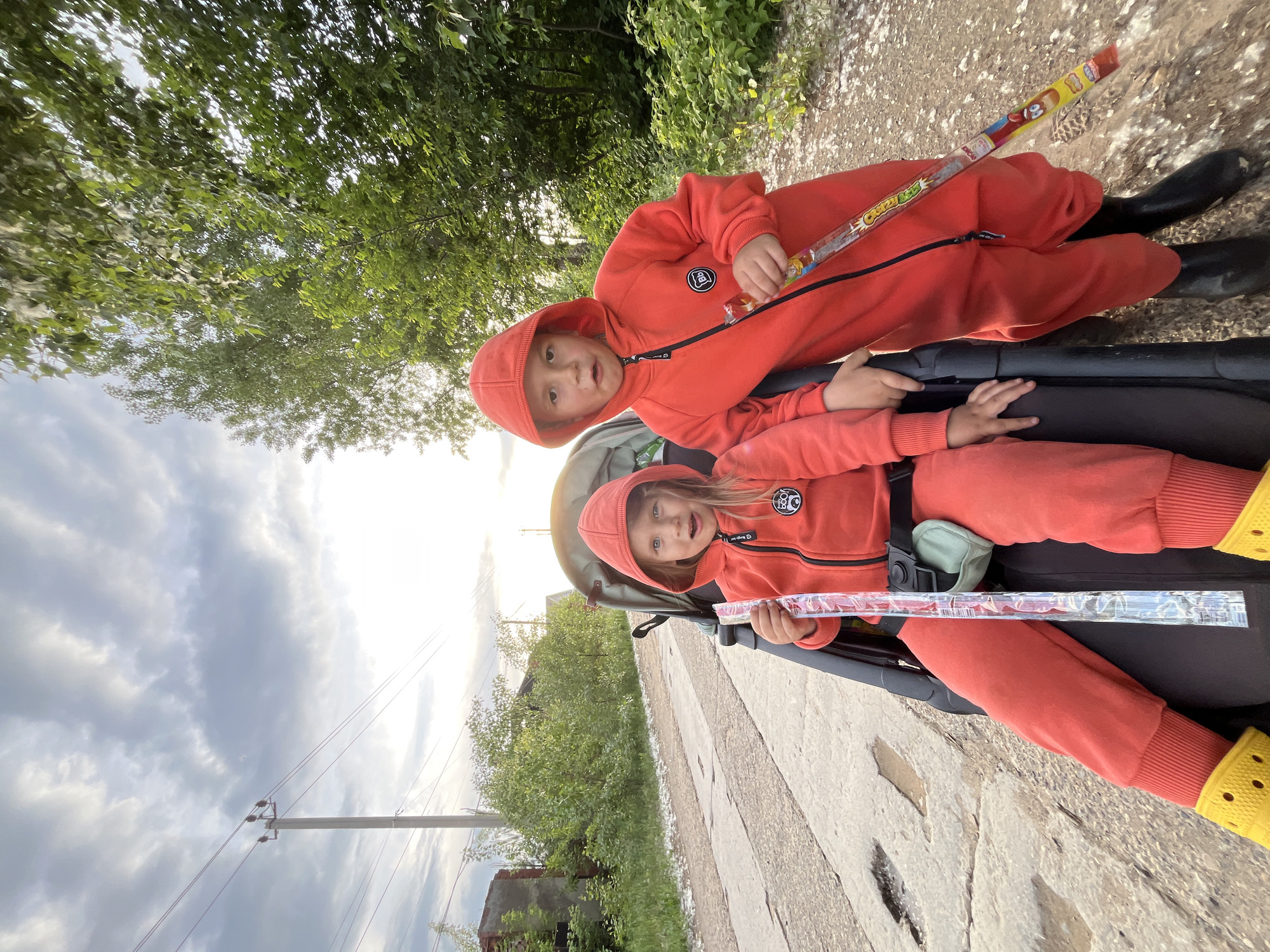 Two children wearing orange hooded jumpsuits are sitting in a stroller, each holding a clear plastic candy tube. One child is in the upper seat, and the other is in the lower seat. The stroller is on a concrete surface, potentially a sidewalk or a crosswalk. There are trees and vegetation visible in the background, along with some buildings and a utility pole with power lines. The sky is cloudy. The location is likely the city of Chaikovsky, Russia.