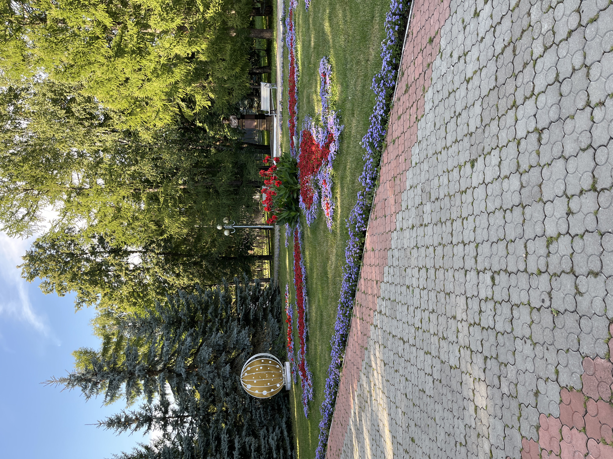 The image depicts a park scene on a sunny day in Almetyevsk, Russia. In the foreground, a paved walkway made of interlocking gray and pink pavers leads into the park. Beyond the walkway, a meticulously maintained flower bed features a vibrant display of red and purple flowers arranged in geometric patterns on a green lawn. A large, spherical art installation with a gold-colored, patterned surface stands prominently to the left of the flower beds. In the middle ground, a statue of a bust is visible, accompanied by a white park bench. The background is filled with lush green trees, including several tall evergreen trees on the left and deciduous trees further back. The sky is blue with a few wispy clouds. The overall impression is one of a well-kept public space designed for relaxation and enjoyment.