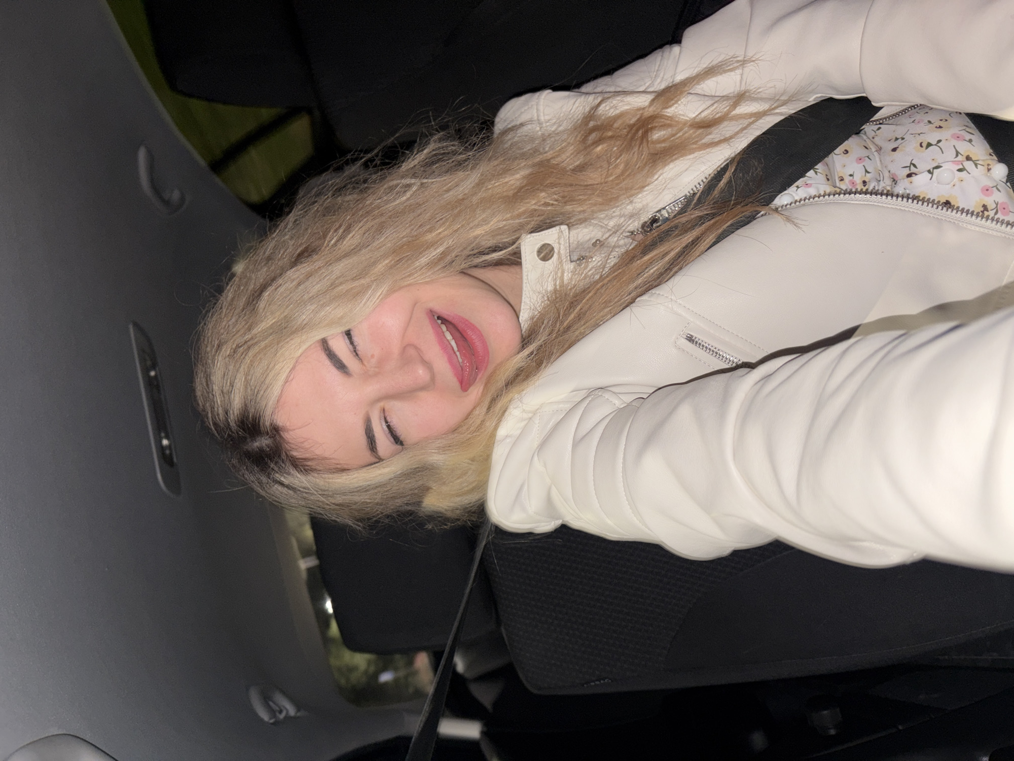 A young woman with blonde hair lies sideways in a car, possibly in Budva, Montenegro, though there's no definitive landmark confirming the location. She wears a white leather jacket over a floral-patterned top and a seatbelt. The car's interior is visible, including the roof, door, and seat. The background suggests a dark outdoor environment.