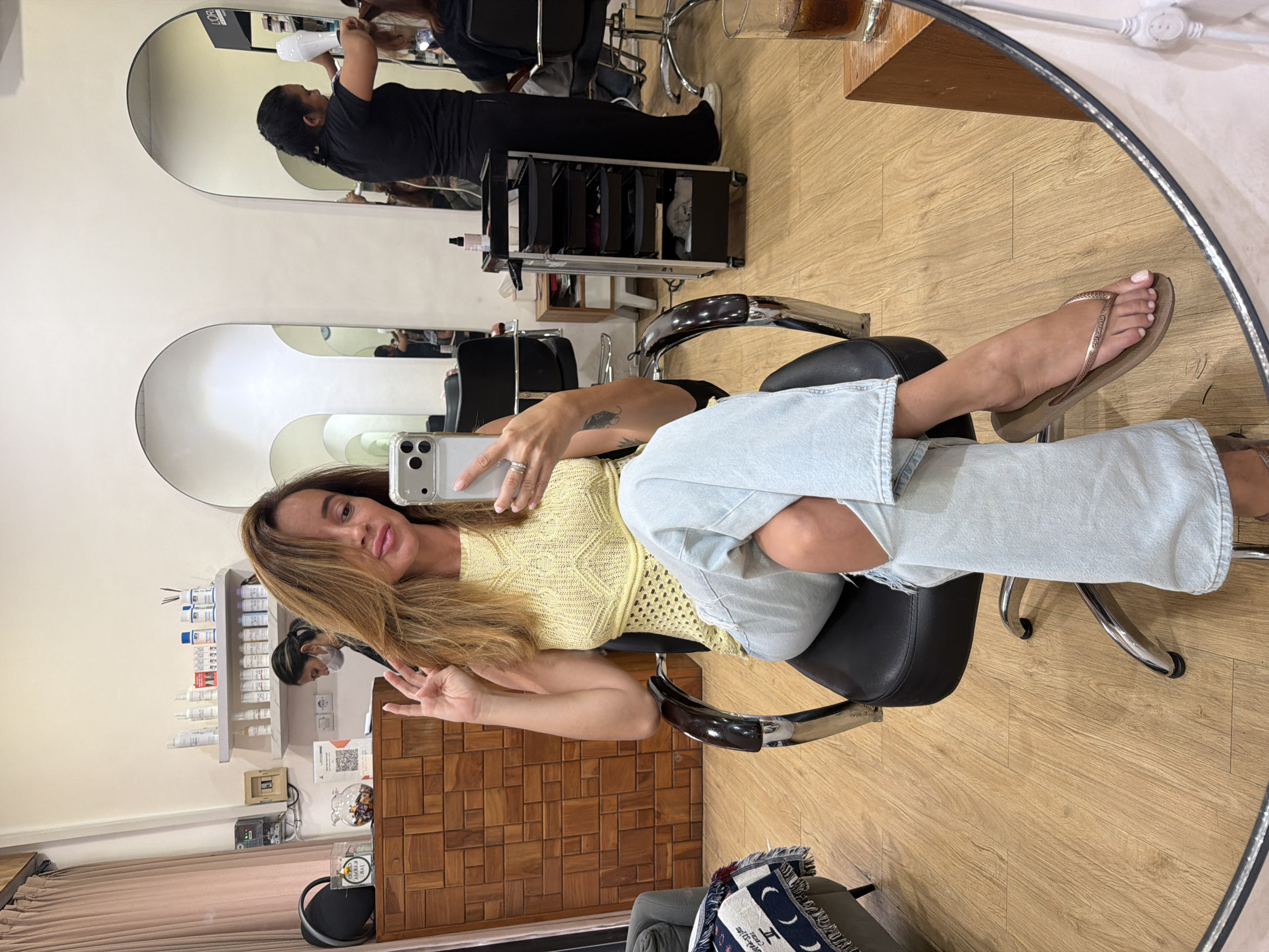 In North Kuta, Indonesia, a woman with long, light brown hair and wearing a yellow knitted top and light blue ripped jeans sits in a black salon chair, holding a smartphone to take a mirror selfie. She is positioned in the foreground, her right hand raised in a peace sign. Behind her, a salon environment is visible, with multiple styling stations and mirrors. In the background, two hairdressers are attending to clients, one with a hairdryer. Shelves stocked with various hair care products line the walls. The floor is light-colored wood, and a wooden cabinet with a textured finish is present to the left. A glass with a dark liquid sits on a wooden surface in the right foreground.