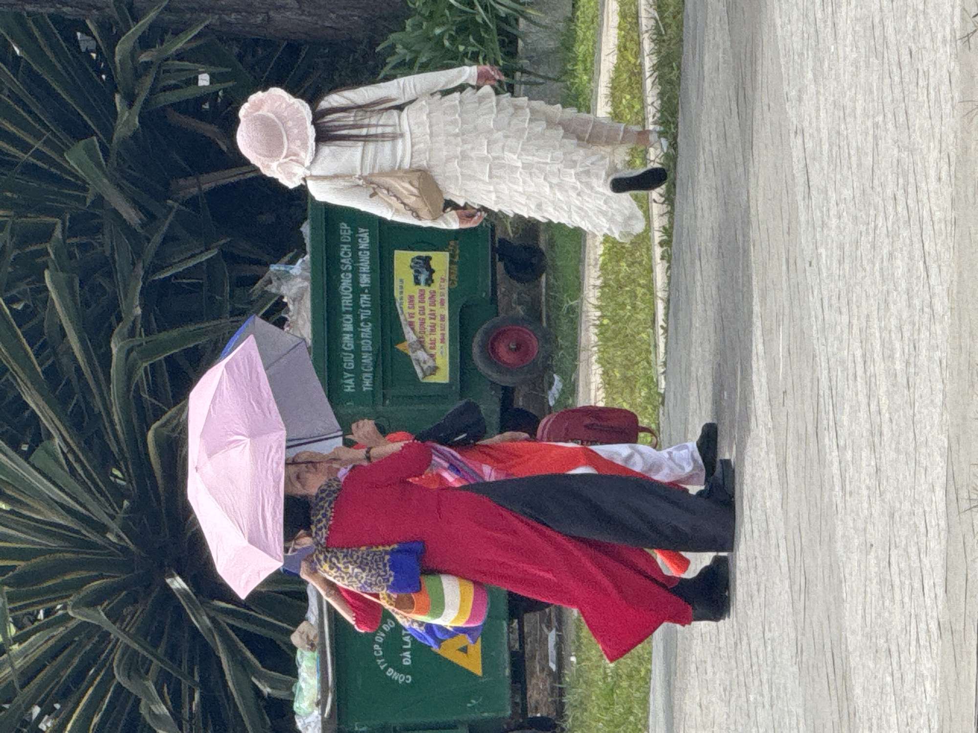 In Da Lat, Vietnam, a group of people are walking outdoors. In the foreground, two individuals are partially visible, one wearing a long, flowing red dress over dark pants and the other in white attire with a ruffled skirt. Both are walking on a paved surface. Behind them, a large green dumpster is visible, with text in Vietnamese printed on its side. The dumpster is surrounded by lush green foliage. Further in the background, a person in a wide-brimmed, light pink hat is walking away from the camera, dressed in a long, tiered white skirt and a light-colored top. One of the individuals in the foreground is holding a pink umbrella over themselves and another person, shielding them from the sun. The environment appears to be a street or public area in Da Lat, indicated by the typical Vietnamese text on the dumpster and the presence of dense tropical vegetation.