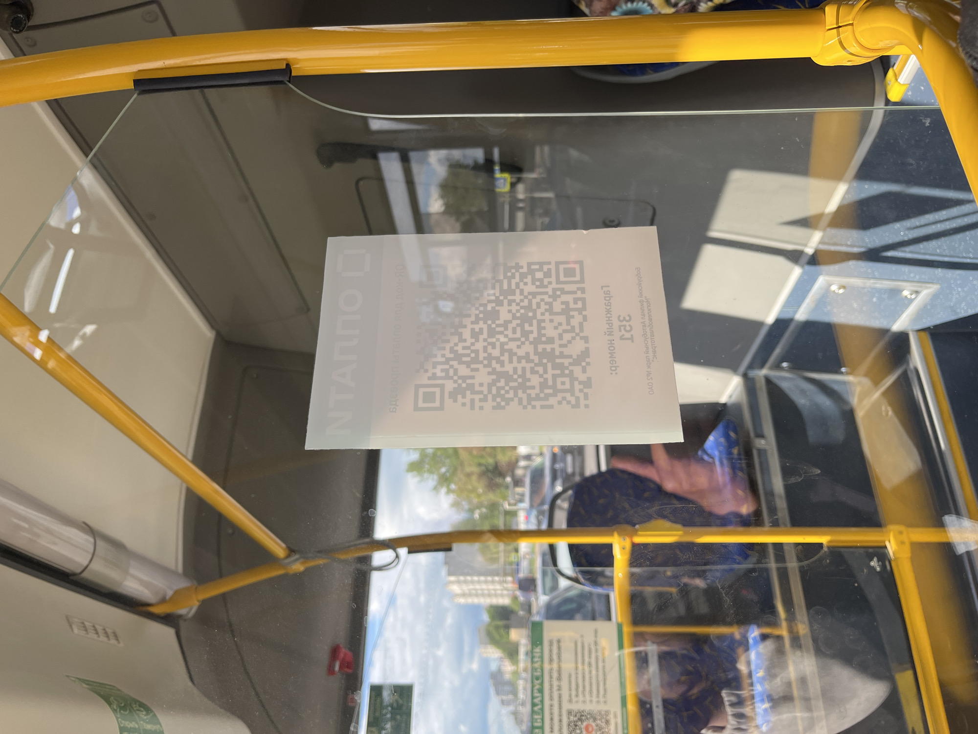 The image shows the interior of a bus, likely in Babruysk, Belarus, as indicated by text on a green poster visible on the left side of the image. The poster, in Belarusian, contains information about "БЕЛАРУСБАНК" and a QR code for payments or information. A prominent white sticker with a QR code and Cyrillic text in white is affixed to the glass partition in the foreground, partially obscuring the view of the bus interior. The Cyrillic text above the QR code appears to be "НТАППО" and below it, in a reversed orientation, is text that reads "ТАРИФ 321". The bus interior features yellow grab poles and blue and grey seats. The overall lighting suggests daytime.