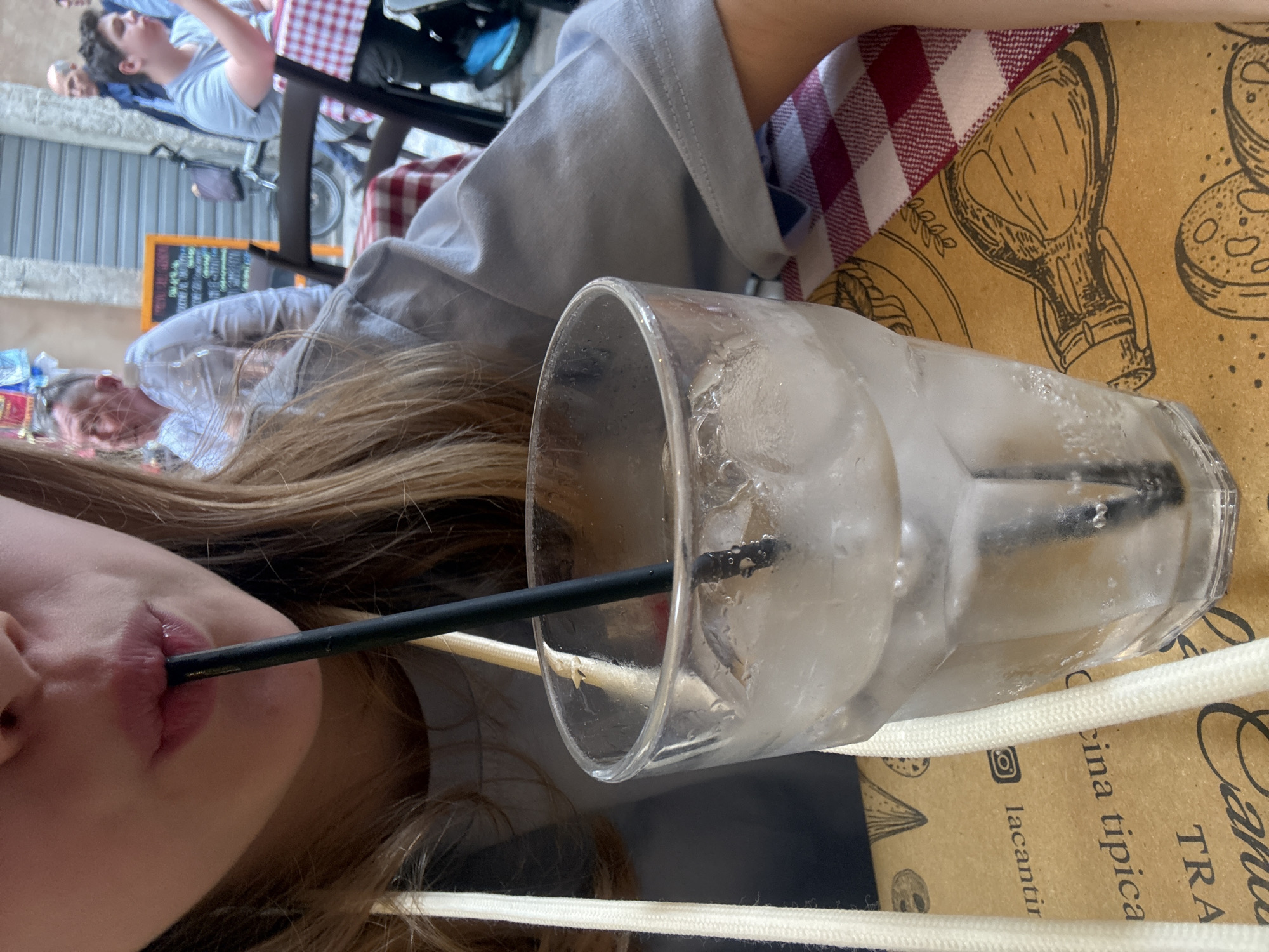 An eye-level shot captures a person drinking from a tall, clear glass filled with ice and a black straw in the foreground. Only the person's lower face, showing their lips around the straw, and long, light brown hair are visible. The glass rests on a brown paper placemat adorned with illustrations of food items, including pizza slices, pasta shapes, and an amphora or jug. A red and white checkered tablecloth is visible beneath the paper placemat.
In the midground, another person, wearing a light blue t-shirt, sits at a table, partially obscured. Behind them, a third individual, an older male, is vaguely visible. The background reveals an outdoor setting, likely a street or piazza in a European city, consistent with Rome. Buildings with closed roller shutters and a parked bicycle can be seen. An orange chalkboard sign, probably a menu, stands further in the background.
The bright lighting suggests it is daytime, possibly a sunny day. The frosted glass implies a cold beverage, offering refreshment. The checkered tablecloth and illustrated placemat with text like "cina tipica" (likely part of "cucina tipica" for typical cuisine) and "lacantir" (possibly "La Cantina" with an Instagram logo) strongly indicate an Italian trattoria or cafe. The overall scene depicts a casual dining experience outdoors.