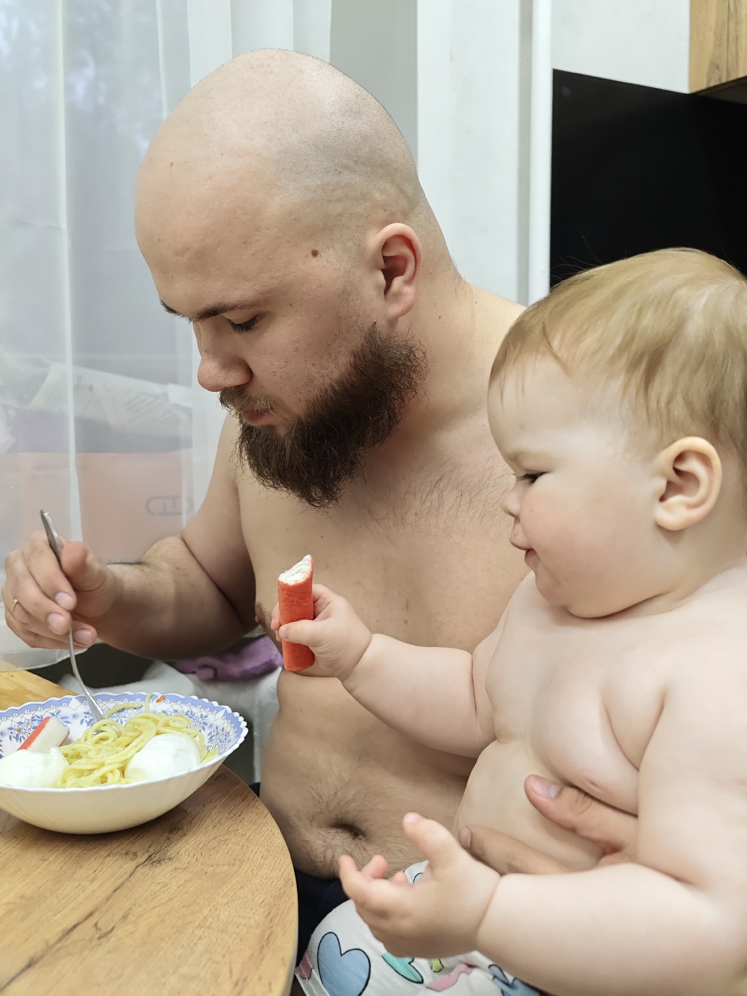 A bald, bearded man sits shirtless at a wooden table, holding a fork and eating pasta and what appears to be boiled eggs from a bowl with a blue floral pattern. He is also holding a toddler, who is also shirtless and holding a crab stick. The location is not discernible from the image.