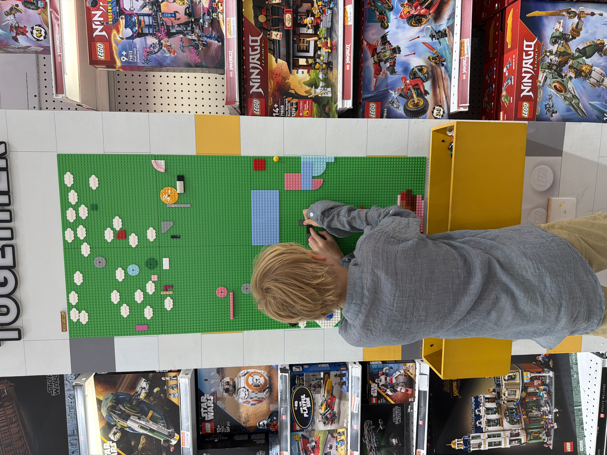 A boy is building Legos on a green baseplate that is on a shelf stocked with Lego sets. The boy is wearing a grey shirt and tan shorts. The Lego sets are from the Star Wars, Ninjago, and Icons product lines. The sets are on a shelf in a retail store.
