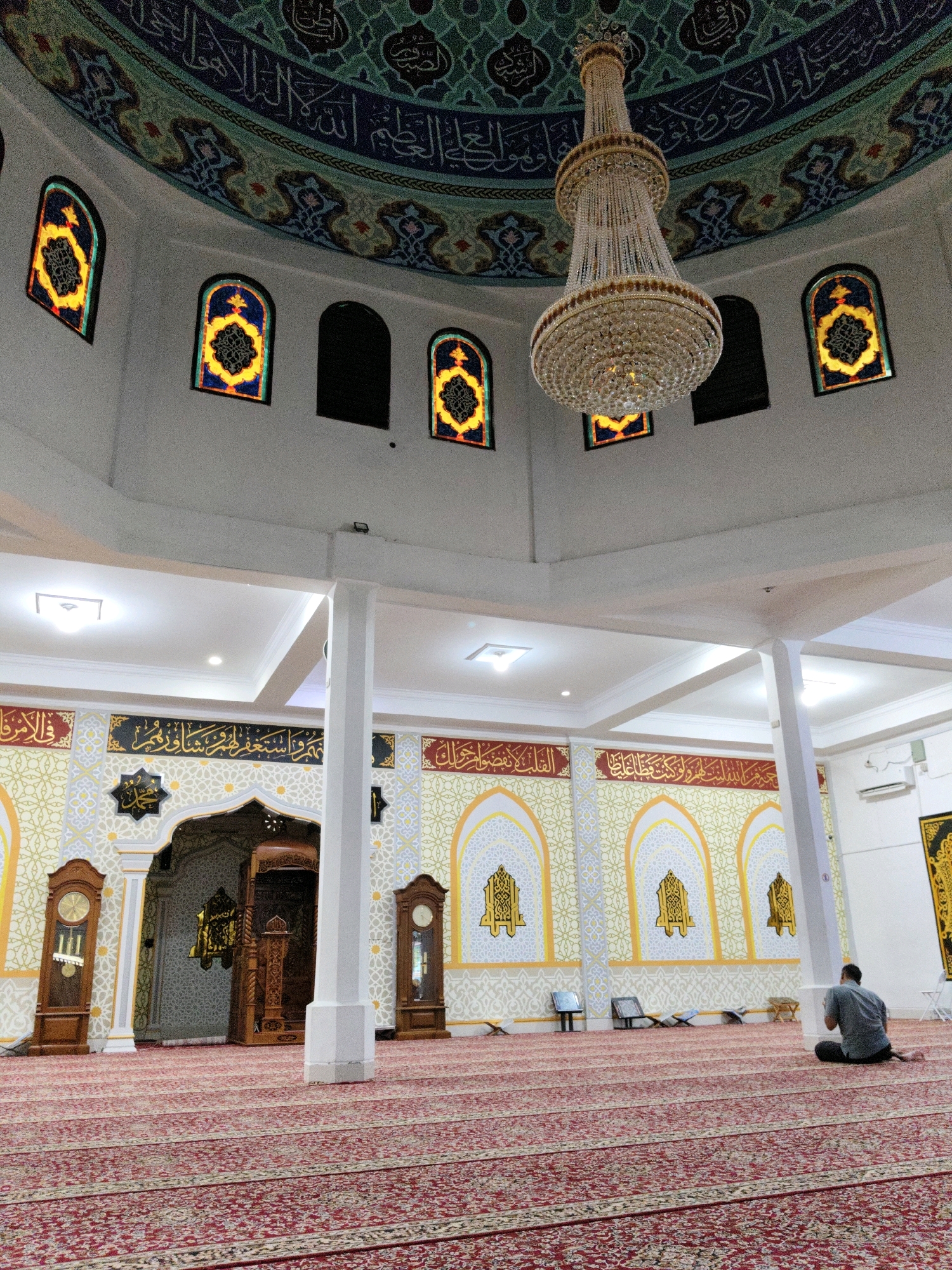 This image depicts the interior of a mosque in Lhokseumawe, Indonesia. The scene is dominated by a large, ornate chandelier hanging from a domed ceiling decorated with Islamic calligraphy and intricate patterns in shades of blue, green, and white. Several arched stained-glass windows with yellow and blue geometric designs are visible along the upper walls.
The mosque features a red carpet with a repeating pattern of beige and dark red motifs, covering the floor. White pillars support the ceiling. On the left side of the image, two tall grandfather clocks flank an ornate wooden entrance to the prayer area. The walls are adorned with decorative panels featuring yellow and white geometric patterns and Arabic script.
In the foreground, a man wearing a grey t-shirt and dark pants is seated on the carpet, facing away from the camera, in a prayer posture. He appears to be engaged in prayer or supplication. Various objects, including what seem to be prayer mats or books, are placed on the floor near the back wall. The lighting is a mix of natural light filtering through the windows and artificial illumination from ceiling fixtures. The overall atmosphere is one of serenity and devotion.