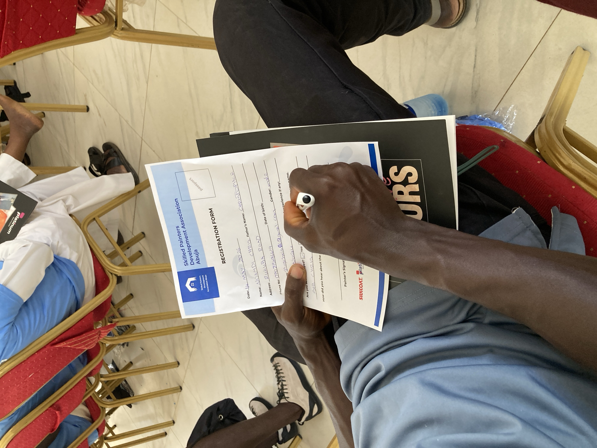 A person is filling out a registration form for the "Skilled Painters Development Association Abuja." The form includes fields for the date (4 MAY 2026), the applicant's name (Nwunendi Chris), father's name (Christopher), address (Rukuba Road), phone number (07087496619), email (ploychris06@gmail.com), date of birth, gender (male), and previous experience. The person is signing the form as "Sir Chris." The address on the form, Rukuba Road, is located in the city of Jos, Nigeria.