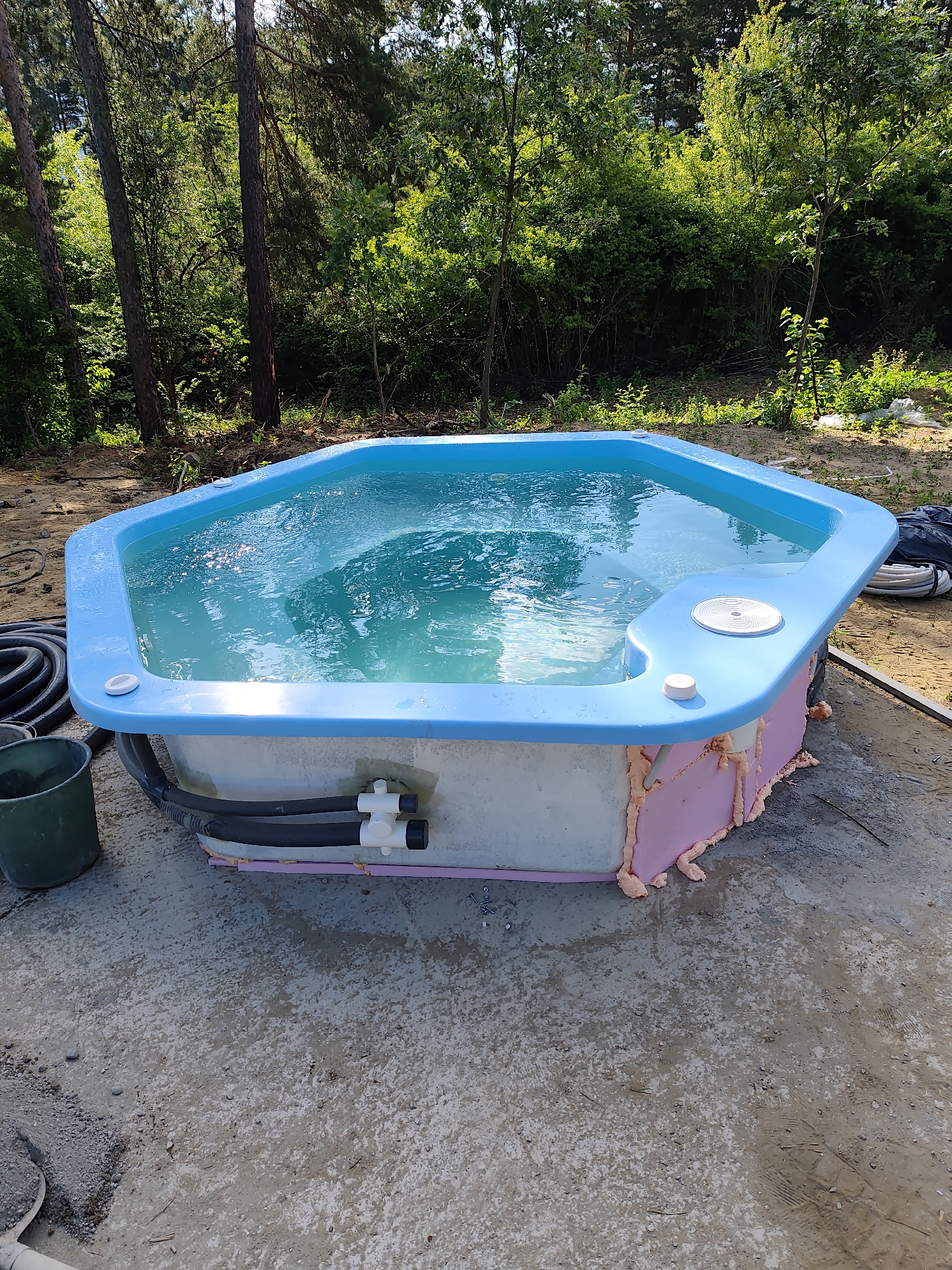 A partially installed, hexagonal blue-rimmed hot tub filled with turquoise water sits on a gray concrete pad surrounded by a wooded area with tall pine trees and dense green foliage. The exterior of the hot tub is insulated with pink foam. Black hoses and a white plumbing fixture are attached to the side. A green bucket and coiled black hose are to the left of the hot tub. The ground around the hot tub is a mix of concrete, sand, and some scattered debris. The scene appears to be in Kladnitsa, Bulgaria.