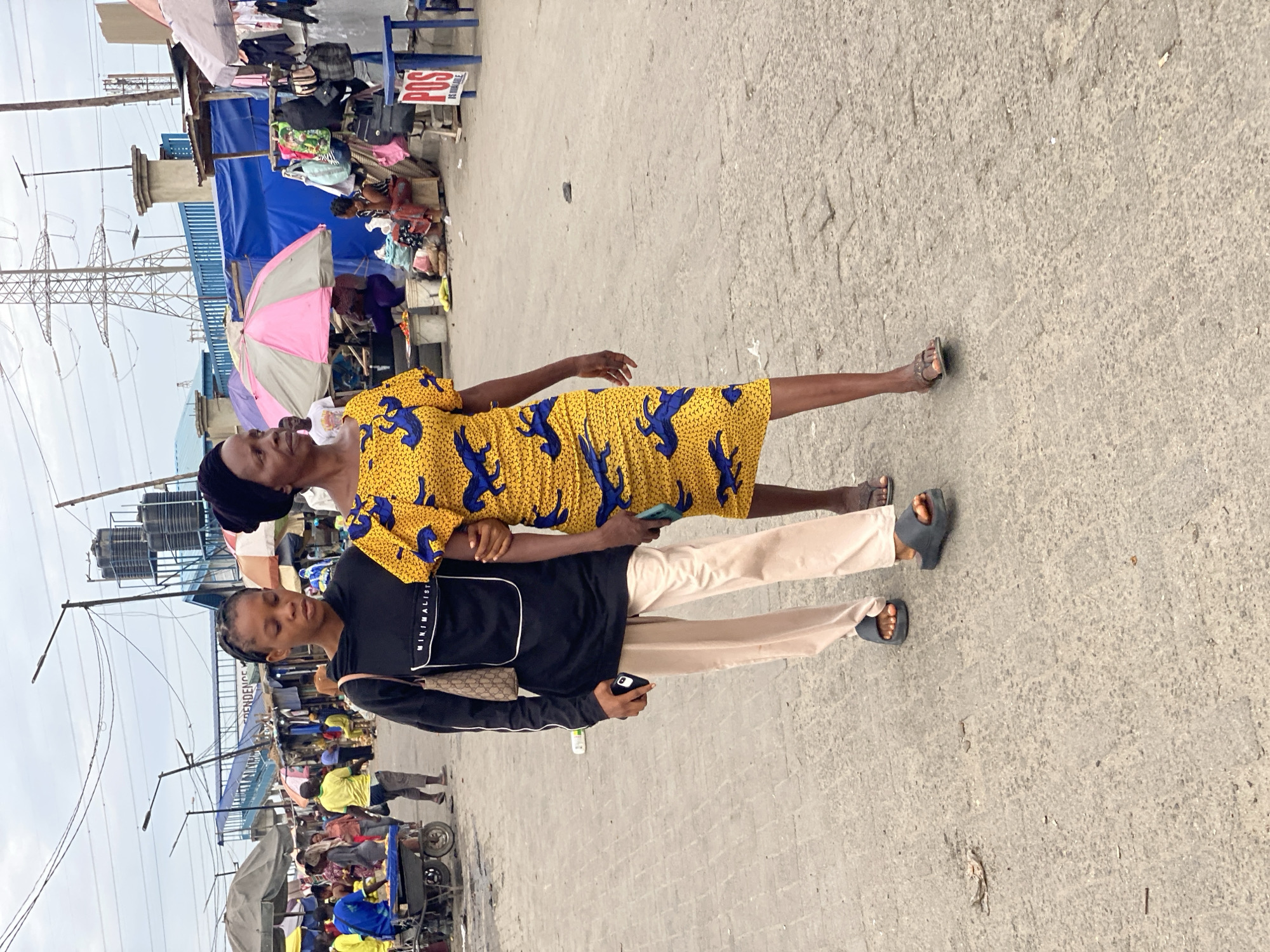 The image shows two women in a street setting, presumably in Lagos, Nigeria, given the presence of street vendors, pedestrian traffic, and an informal market environment.
One woman is wearing a yellow dress with a blue pattern. The other woman is dressed in a black long-sleeved shirt with a white logo on the front and light-colored trousers. She carries a purse and holds a cell phone in her left hand. Both women are wearing sandals. The first woman has her arm around the other.
Behind them, an outdoor market with numerous vendors and customers under umbrellas and temporary canopies is visible. A blue structure with a “POS AVAILABLE” sign is visible at a stall. Power lines and utility poles are also present. The ground is paved with concrete, with visible cracks and imperfections.