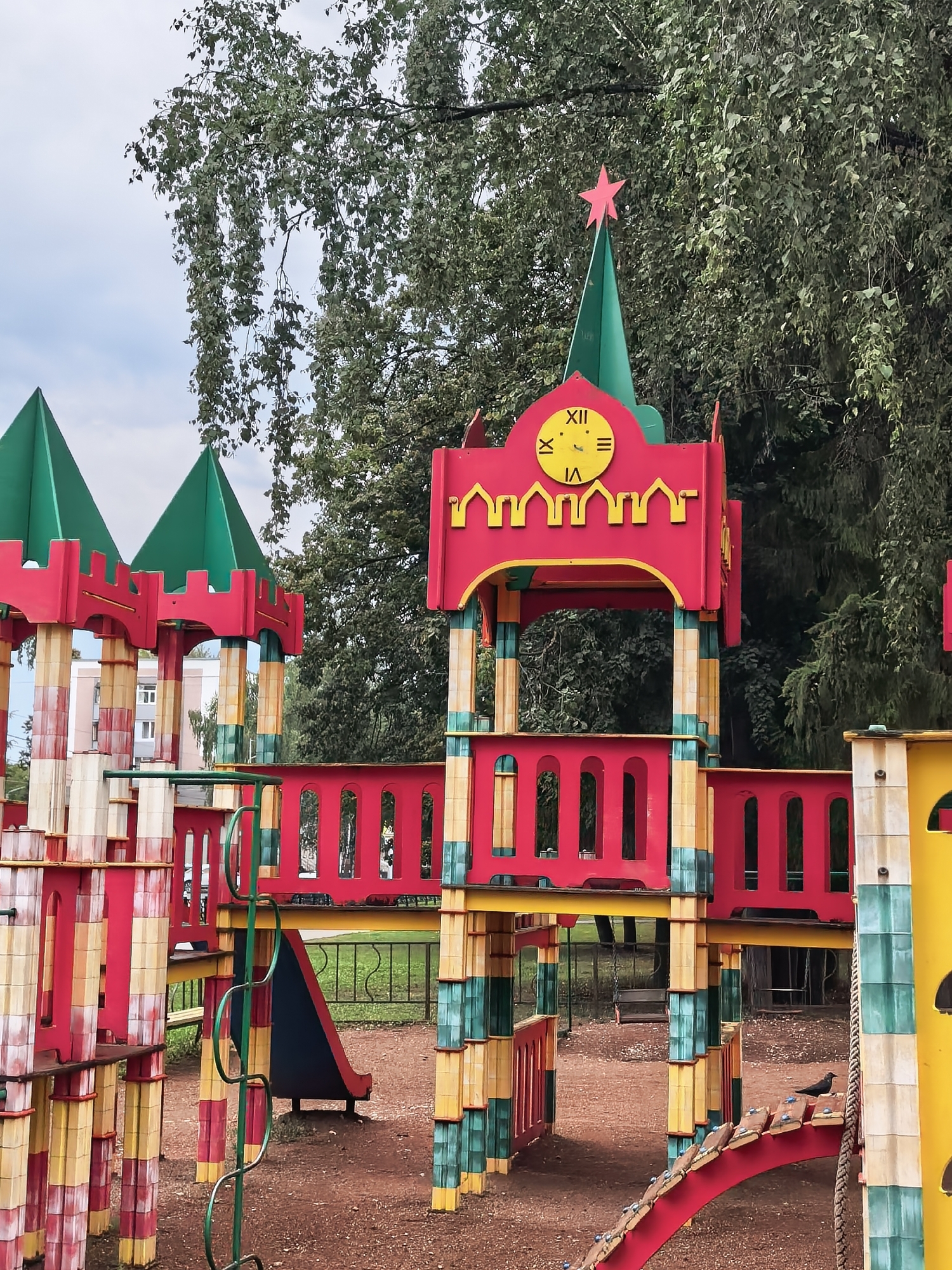 This image depicts a colorful children's playground, possibly located in the city of Sterlitamak, Russia, given the architectural style of the building in the background which resembles Soviet-era apartment blocks. The playground features a large, multi-level structure with red and yellow accents, designed to resemble a castle. It includes turrets topped with green conical roofs and a red star at the apex of one turret. A clock face with Roman numerals is visible on the central tower. The playground equipment is made of brightly colored plastic and metal, including slides, ramps, and climbing structures. The ground surface is covered with reddish-brown gravel. In the background, a large tree with dense foliage partially obscures the sky and a multi-story building. A crow is perched on a red bridge-like ramp in the foreground. The scene is set outdoors under an overcast sky.