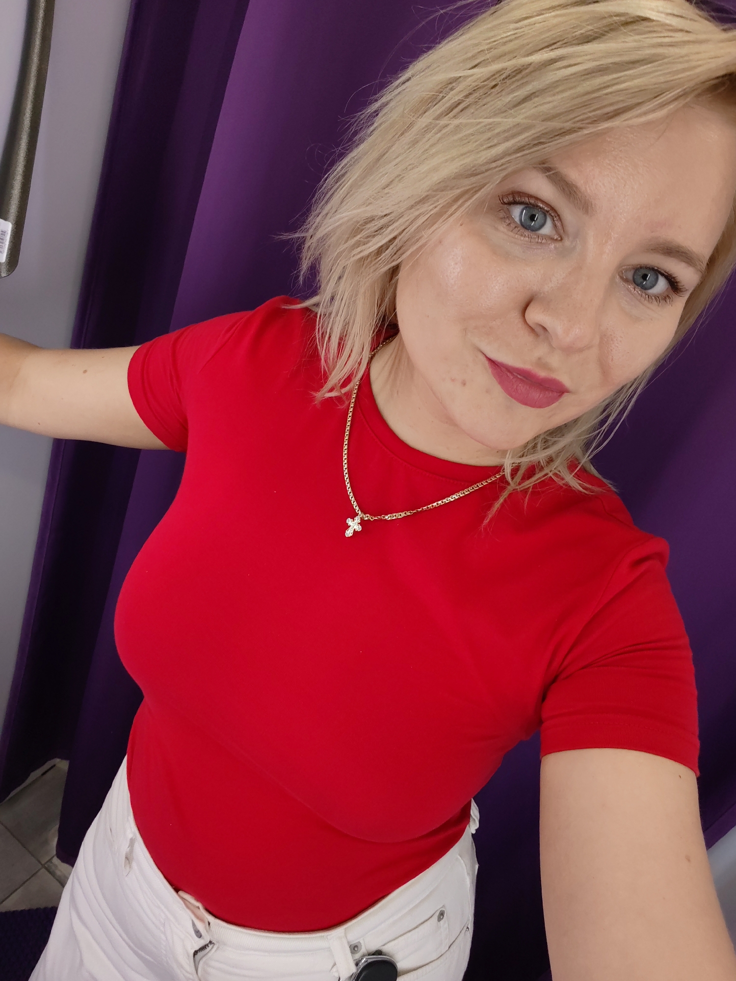 A blonde woman with blue eyes and pink lipstick is taking a selfie in front of a purple curtain. She is wearing a red t-shirt and white pants. She has a gold chain with a cross pendant around her neck.