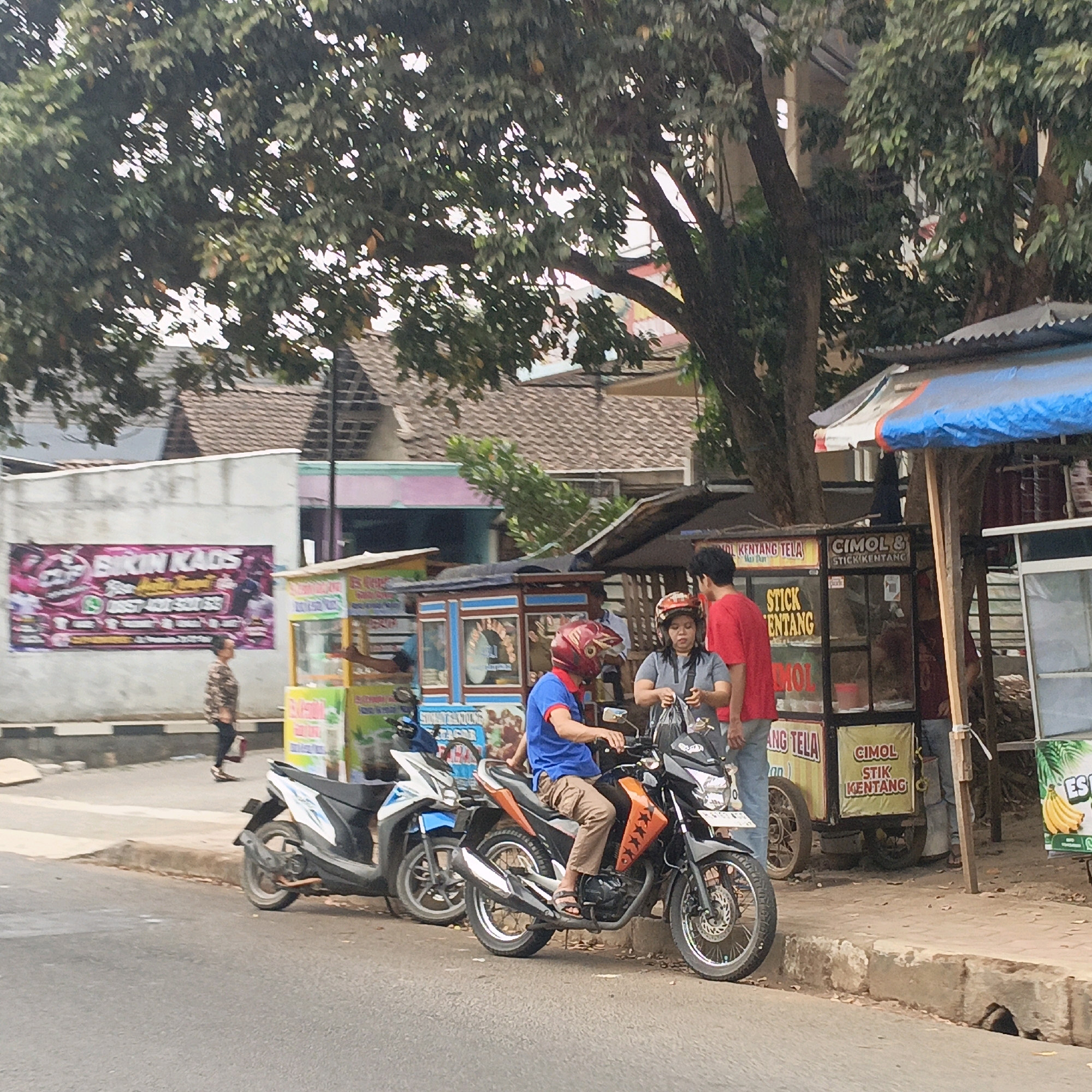 In Salatiga, Indonesia, a street scene unfolds with several individuals and vehicles. A man in a blue shirt and brown pants, wearing a red helmet, is seated on a black and orange motorcycle, positioned on the right side of the frame. Beside him, a woman in a gray shirt and helmet holds a black bag. To their left, another man in a red shirt stands. Behind them, two food stalls are visible, one with a blue and white awning and the other with a blue and red awning. The stalls display signs with text, including "CIMOL & STICK KENTANG". A white and blue scooter is parked to the left of the man on the orange motorcycle. In the background, a large banner with pink and purple text that reads "BIKIN KAOS" is mounted on a wall. A person in a patterned jacket walks along the sidewalk in the mid-ground. The environment features a tree with dense foliage and a tiled-roof building. The scene appears to be captured during daylight.