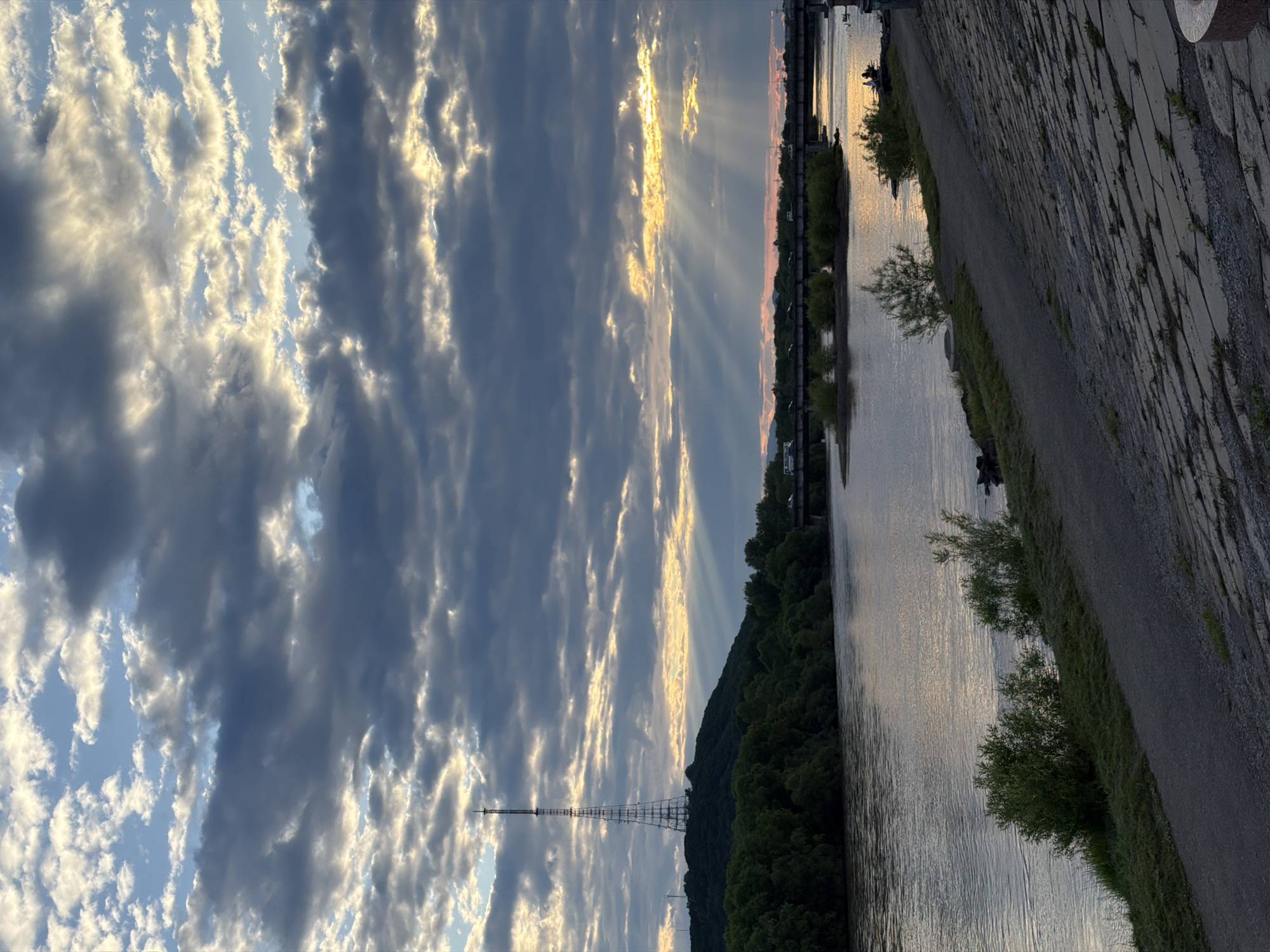 The image depicts a scenic view of the Amur River in Birobidzhan, Russia, at dusk. A prominent transmission tower stands tall on a tree-covered hill on the left side of the frame. The sky is dramatic, with a mix of dark, heavy clouds and patches of bright sunlight breaking through, casting visible rays onto the water. A bridge spans the river in the mid-ground, with cars visible crossing it. The river itself is calm, reflecting the sky. The foreground features a paved walkway and a sloped embankment constructed of stone. Some small trees and vegetation line the riverbank. The overall atmosphere is peaceful and serene, with the last light of the day creating a beautiful natural spectacle.