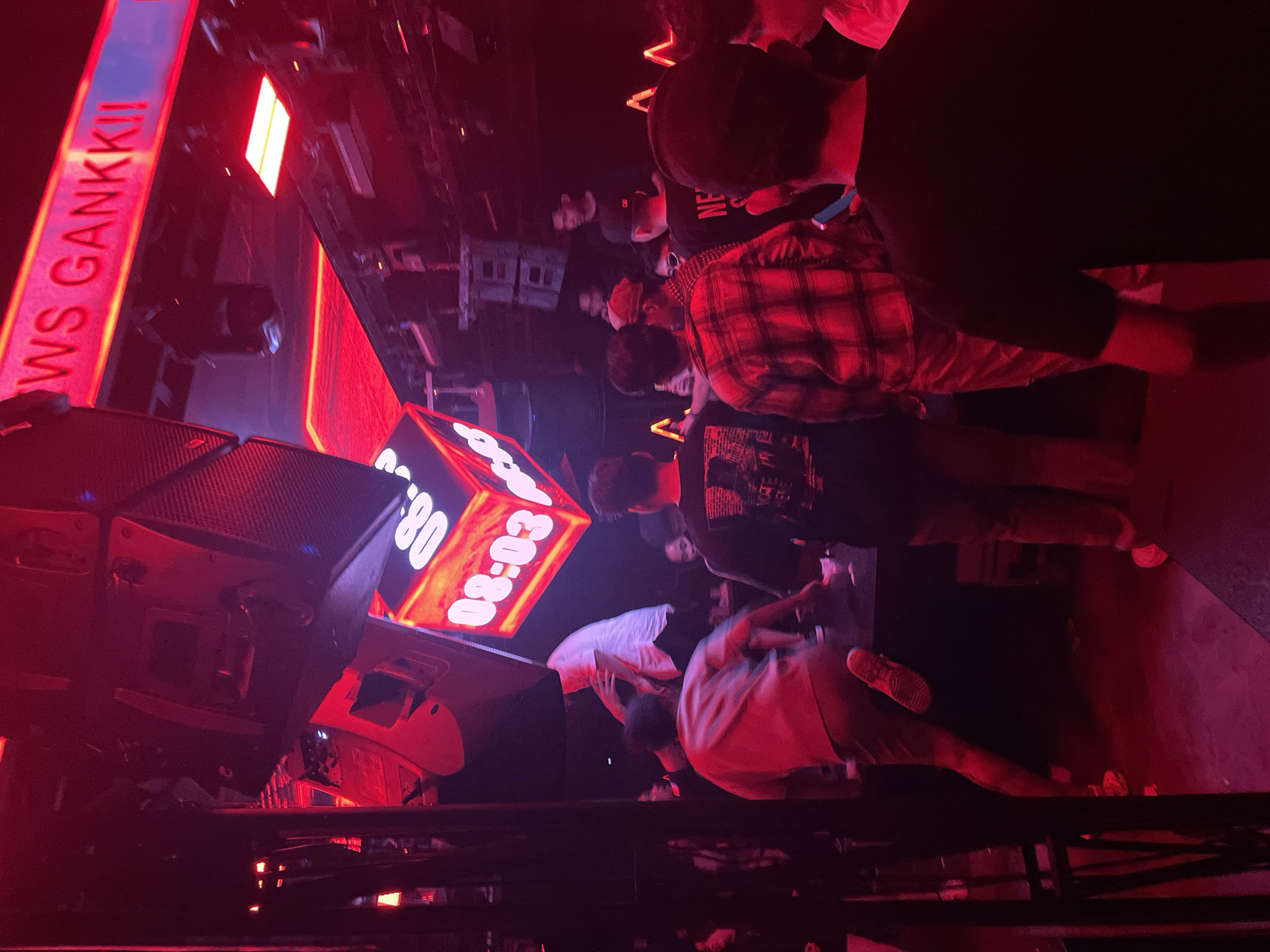 The image depicts the interior of a nightclub in North Jakarta, Indonesia, characterized by vibrant red lighting and a dense crowd. In the foreground, several individuals are visible from behind, engaged in what appears to be a social gathering or dancing. One person in a red and black plaid shirt stands out, while others are dressed in casual attire.
Above the crowd, a large digital clock displays the time "08:03" against a cubic frame with illuminated edges. This prominent clock suggests the time of night or early morning within the establishment. Overhead, large speaker systems and lighting fixtures are mounted, typical of a nightclub environment.
Further back, other patrons are milling about, some appearing to interact with each other. The overall ambiance is energetic and dimly lit, with the dominant red hue creating a nightclub atmosphere. A neon sign with the text "WS GANKKII" is visible at the top of the frame, indicating the name or branding of the venue. The presence of a bar or service area is implied by the presence of people gathered in various parts of the space.