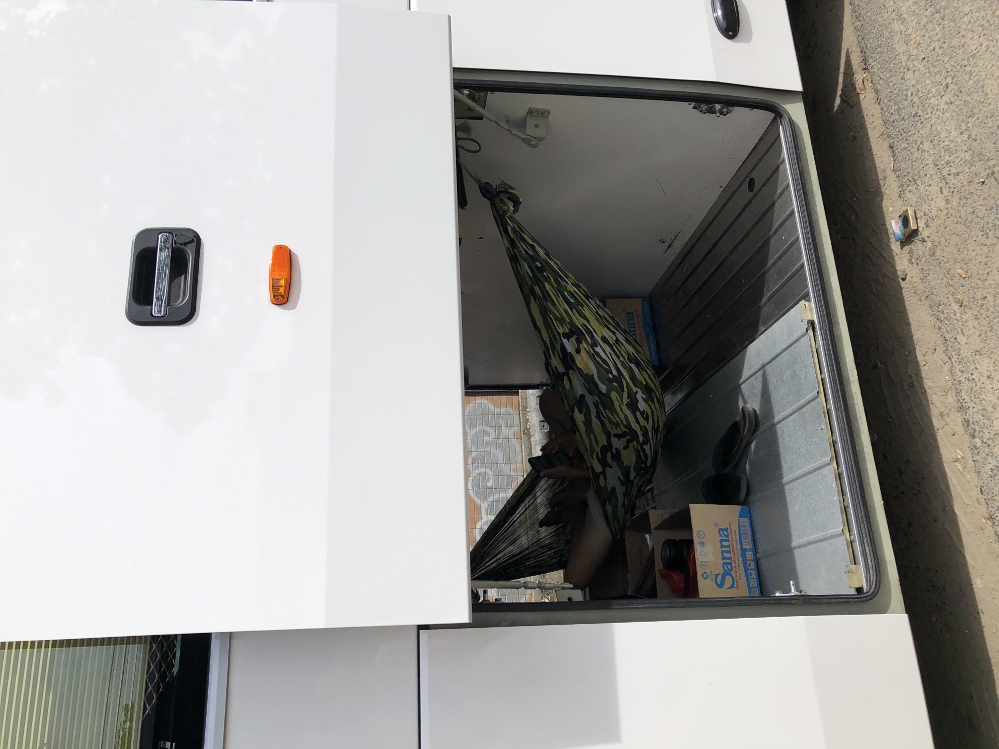A person lies in a camouflage hammock stretched across an open compartment of a white bus. The bus is parked outdoors on a dirt and gravel surface, with a hint of some structure or wall visible in the background with graffiti. The compartment contains a box labeled "Sanna" with water bottles, a pair of flip-flops, and other items. A smartphone is visible in the person's hand. The compartment door above the person is open, revealing the interior of the bus's upper section, which appears to have tinted windows.