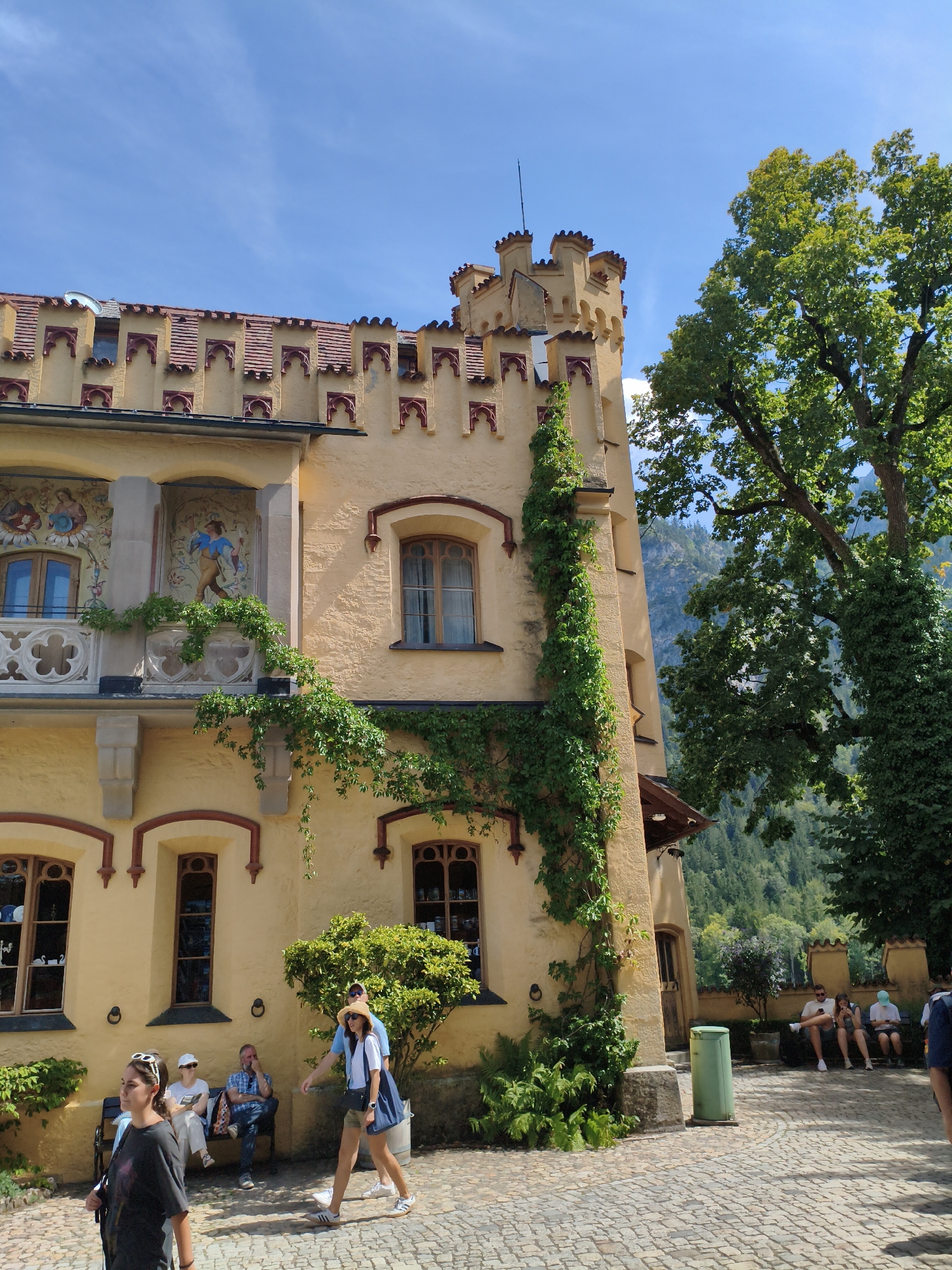 The image depicts Hohenschwangau Castle in Schwangau, Germany, on a sunny day. The castle, painted a pale yellow with red accents along its battlements and window frames, dominates the left side of the frame. Ivy climbs up the side of the building, adding a touch of green to the architecture. Several arched windows, some with decorative glass, are visible. A balcony with an ornate railing features two framed murals, one depicting figures in medieval attire.
In the foreground, a cobblestone courtyard is present. Several people are scattered throughout the scene. On the right, a large, leafy tree provides shade, with its trunk also covered in ivy. Beyond the tree, a mountainous landscape with dense forest is visible under a clear blue sky.
In the courtyard, a group of people are seated on a bench on the left, facing away from the camera. Further to the right, two individuals are walking towards the viewer, one in a blue shirt and straw hat, the other in a dark t-shirt. Another group of people are seated on benches in the middle ground, looking at their phones. A green trash bin is positioned near the seated group. The overall atmosphere is one of a tourist attraction on a pleasant day.