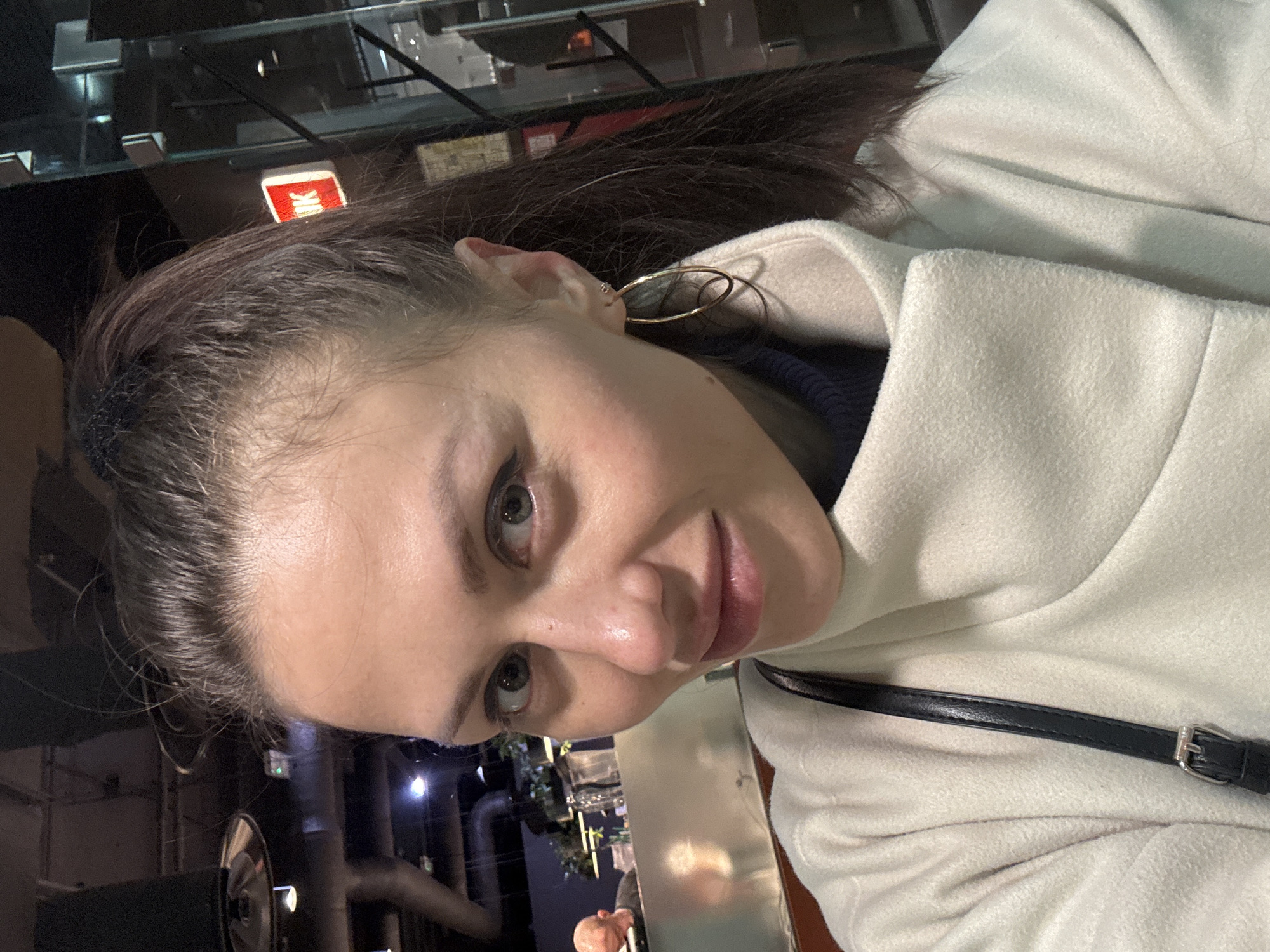 A person with brown hair tied back and wearing gold hoop earrings and a white coat stands in front of what appears to be a window, capturing a selfie. Reflections in the window show elements of a restaurant or bar interior, including a bar area with plants and possibly a pipe structure on the ceiling, suggesting an industrial style decor. The image might be taken in Saint Petersburg, Russia, based on the visible "IK" sign in the window reflection, reminiscent of Cyrillic writing styles.