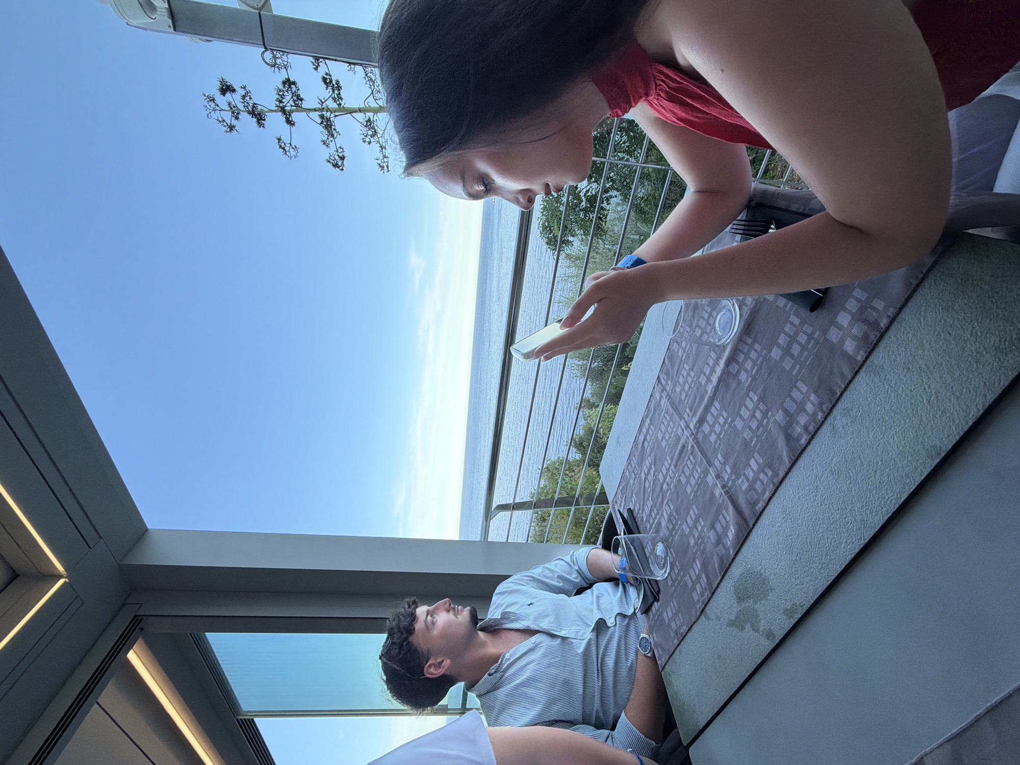 Two individuals are seated at a table on a balcony overlooking the sea in Santa Maria Navarrese, Italy. The man, on the left, is wearing a light blue striped button-down shirt and looking out at the water. He has dark curly hair and is wearing a watch on his left wrist. In front of him, there is a glass and cutlery. The woman, on the right, is wearing a red top and is looking down at her phone. She has long dark hair and is wearing a blue wristband. The table is covered with a patterned cloth. Beyond the balcony, the sea stretches to the horizon under a clear blue sky, with some greenery visible on the lower slopes. The architecture of the building with modern lighting fixtures suggests a contemporary establishment.
